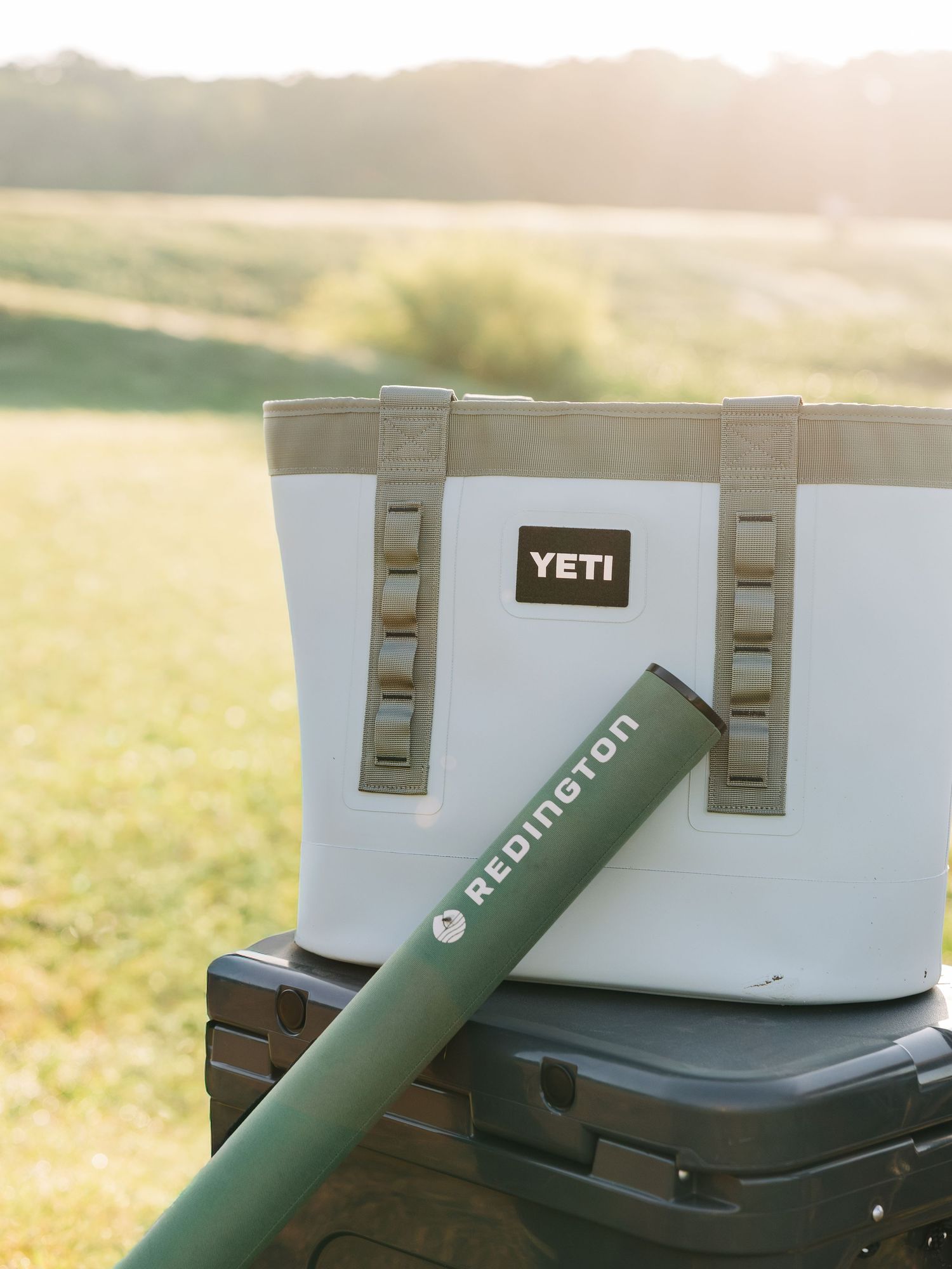 YETI soft cooler bag with tan handles beside a green Redington rod tube outdoors at golden hour.