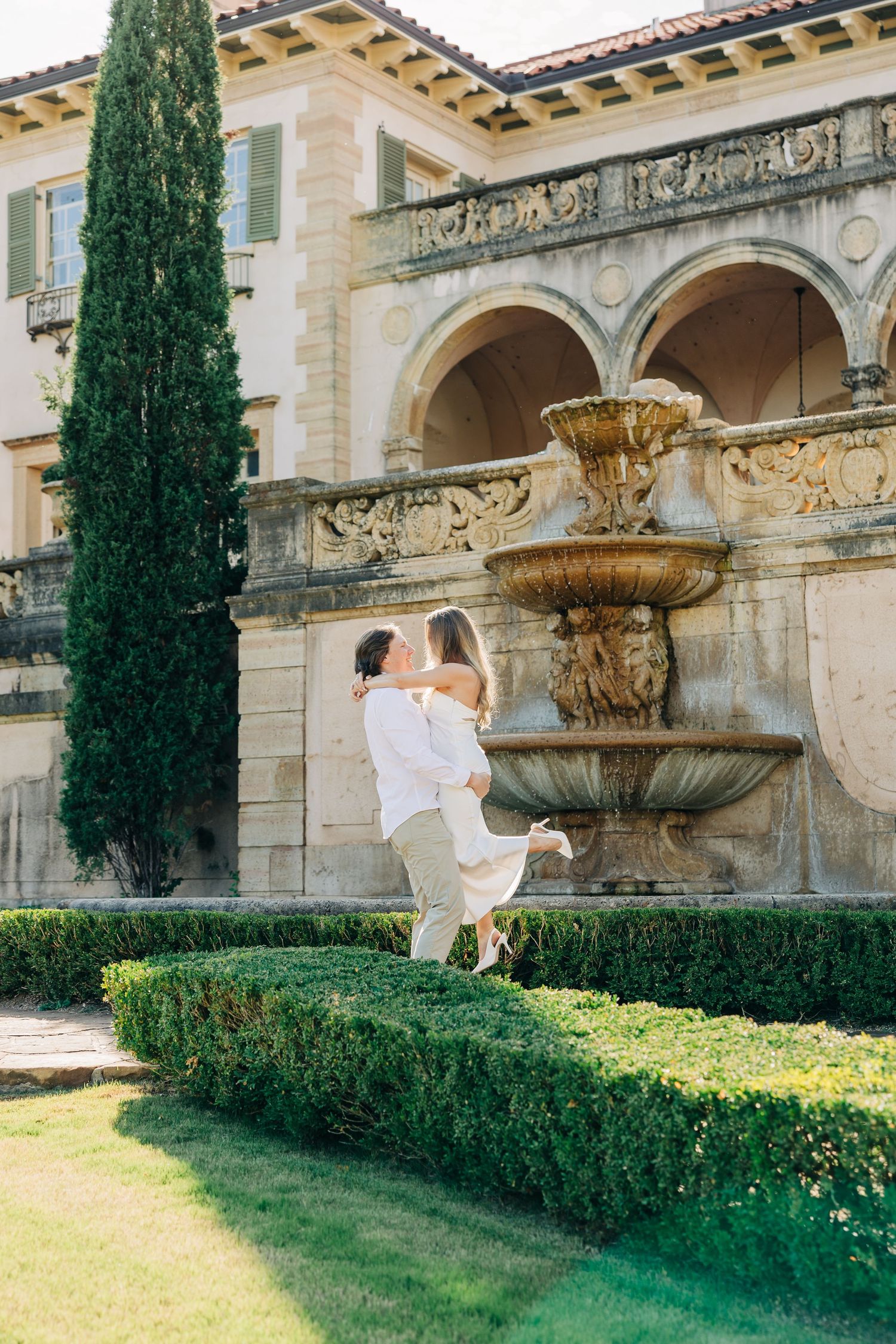 A couple in white wedding attire poses near ornate stone fountains and cypress trees in a manicured garden setting.