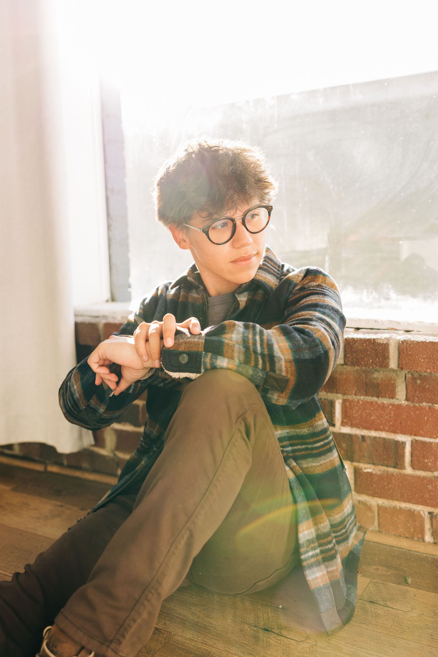 Person in glasses and plaid shirt lounges against brick wall in series of artistic portraits with warm lighting.
