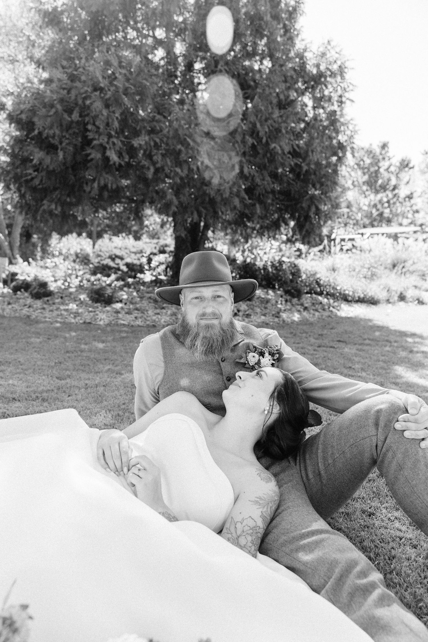 Couple lying on grass in romantic black and white wedding photo with trees in background.