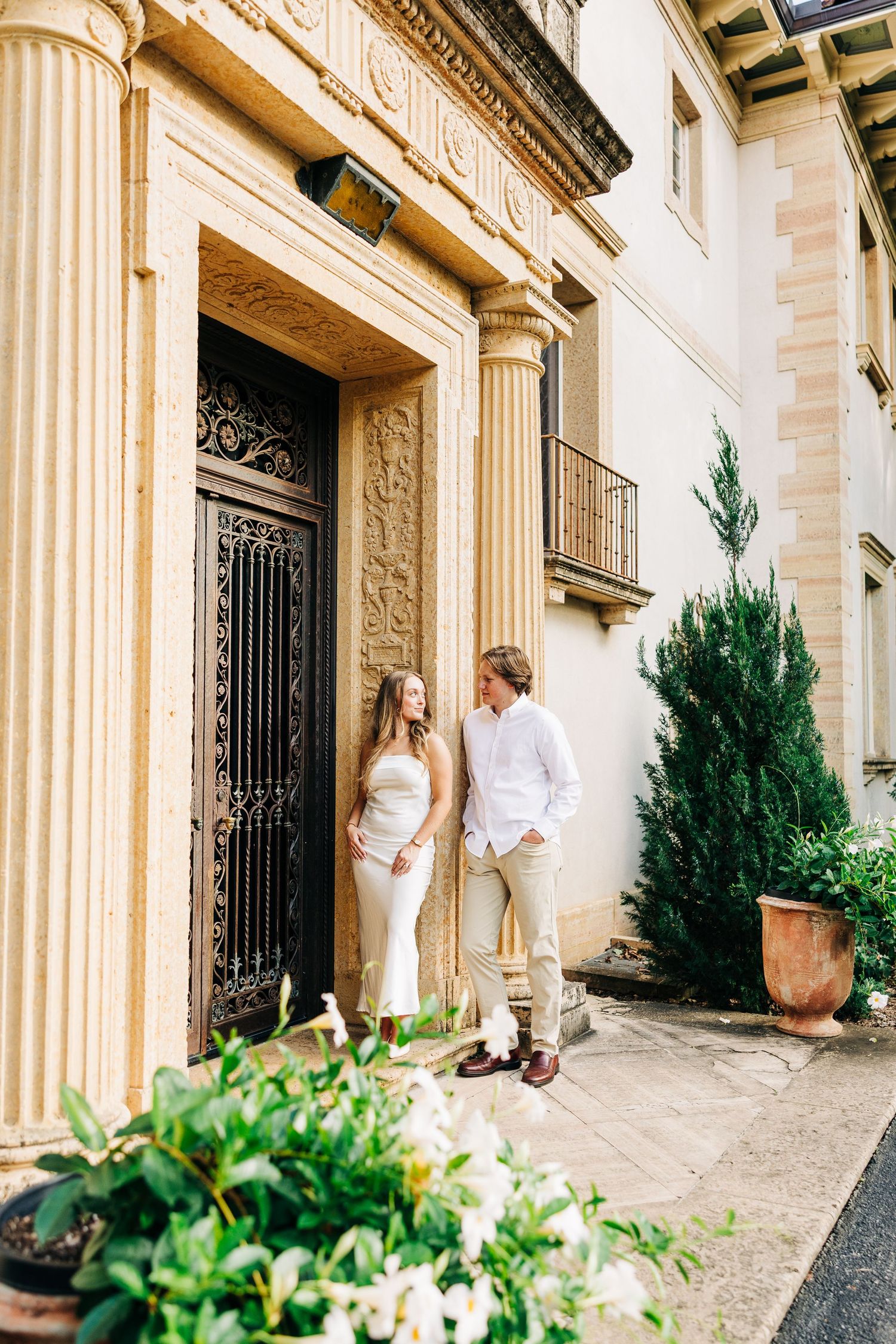 Elegant couple poses against ornate stone columns and greenery at a grand architectural entrance.