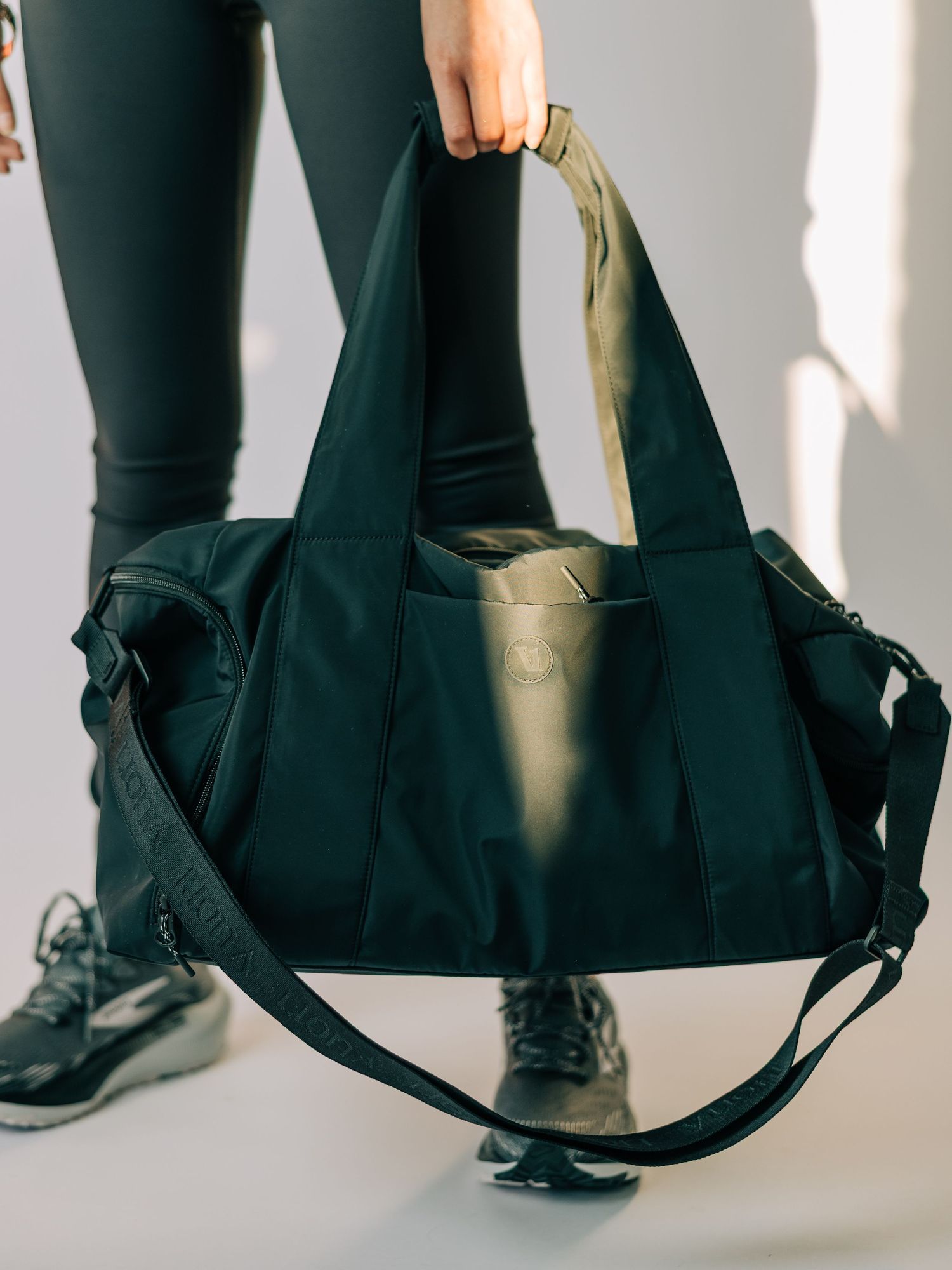 Person holding a black gym duffel bag with gold handles and shoulder strap, wearing dark leggings and sneakers.