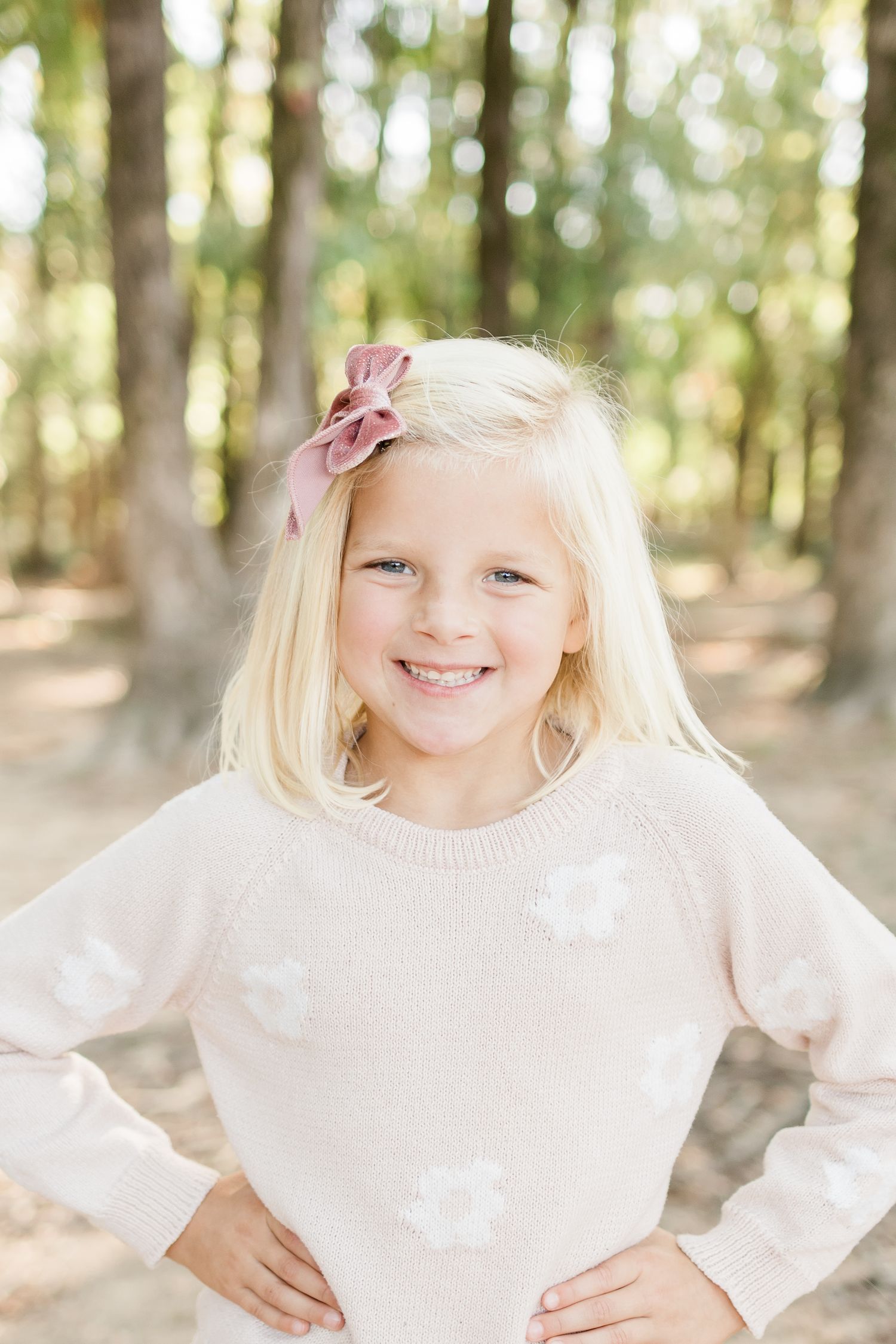 Joyful children in white sweaters pose playfully in a sunlit forest setting with bokeh effect in background.