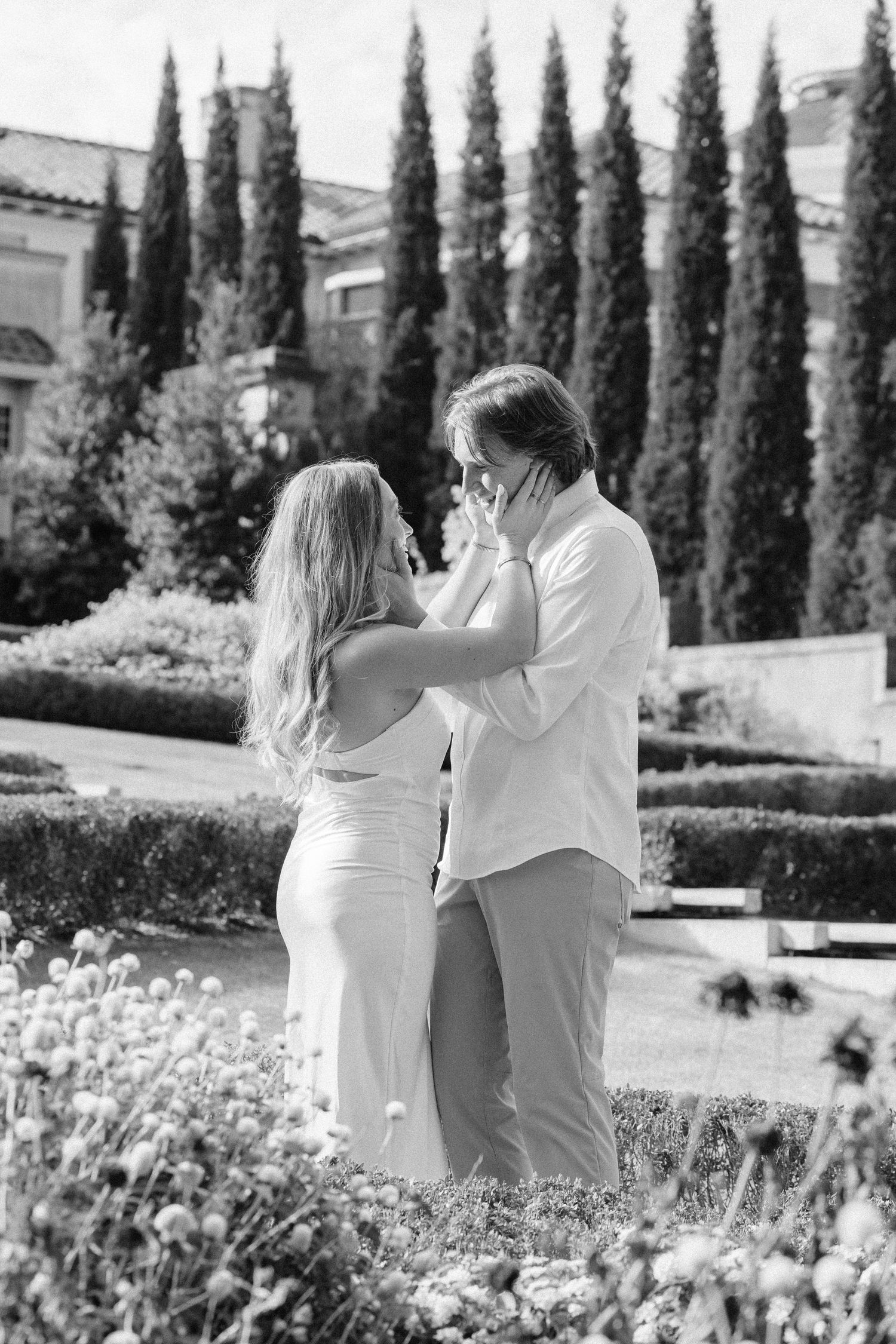 A couple embraces while walking through a garden with tall cypress trees and blooming flowers in black and white.