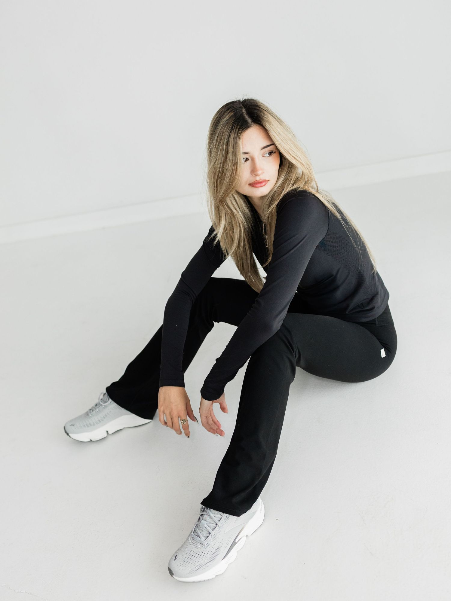 Woman in black athletic outfit and gray sneakers sitting on white floor, showcasing stylish activewear.