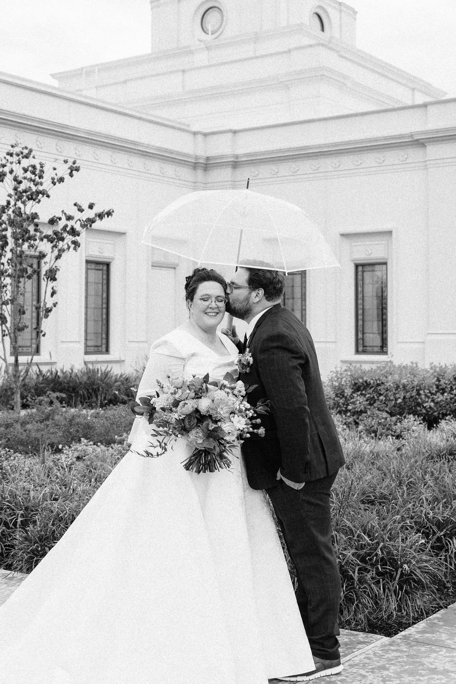 Couple shares an intimate moment under a clear umbrella outside a white church building.