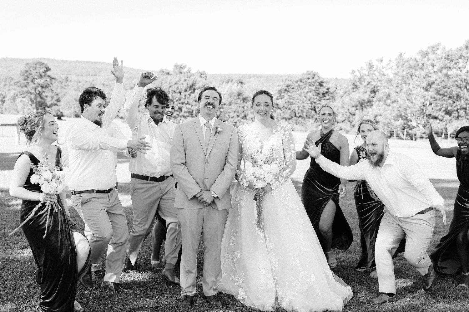 A joyful wedding party in light colored attire poses and dances together in an outdoor garden setting.