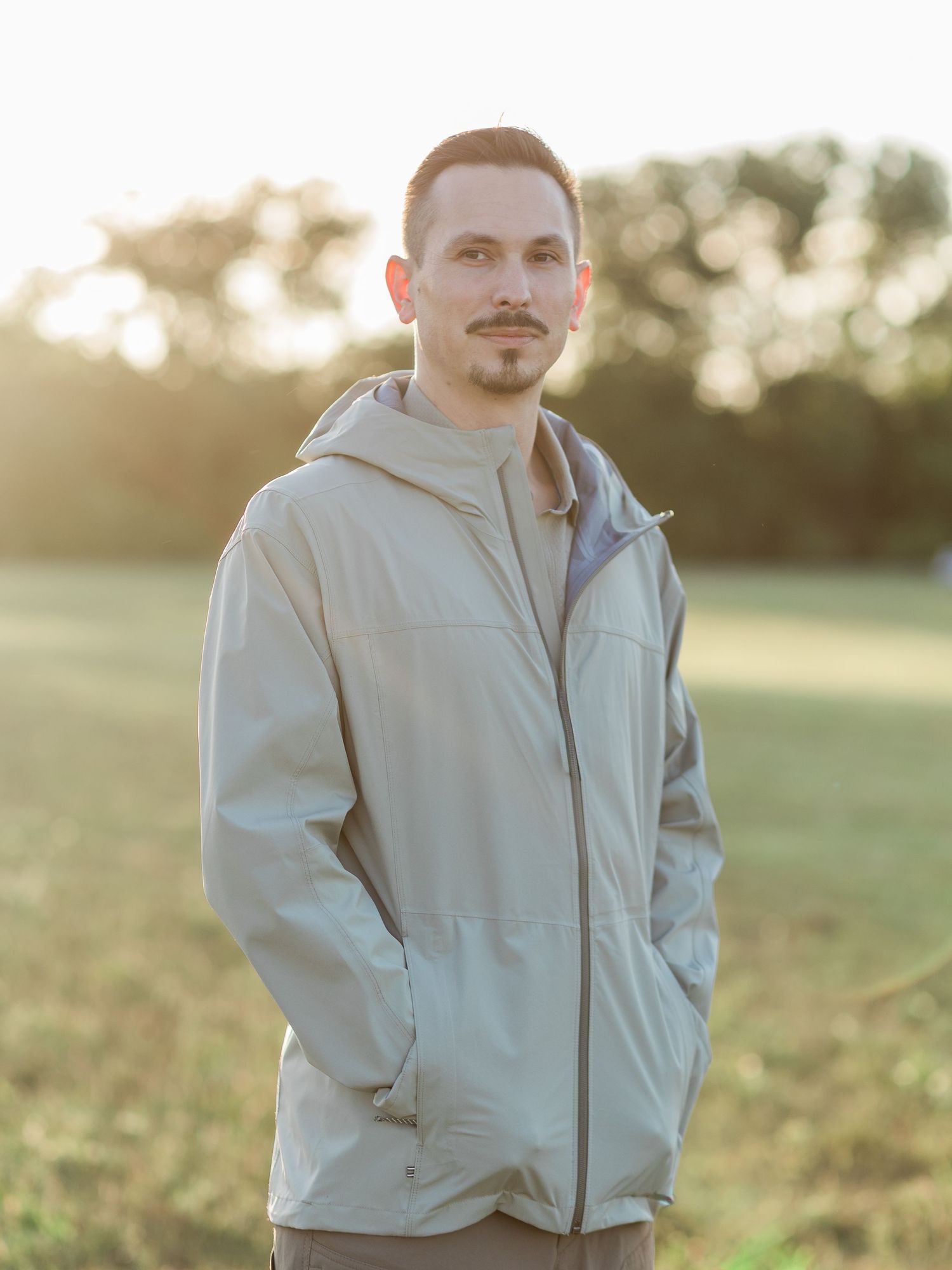 Young man in light gray hooded jacket standing in sunlit field with trees in background.