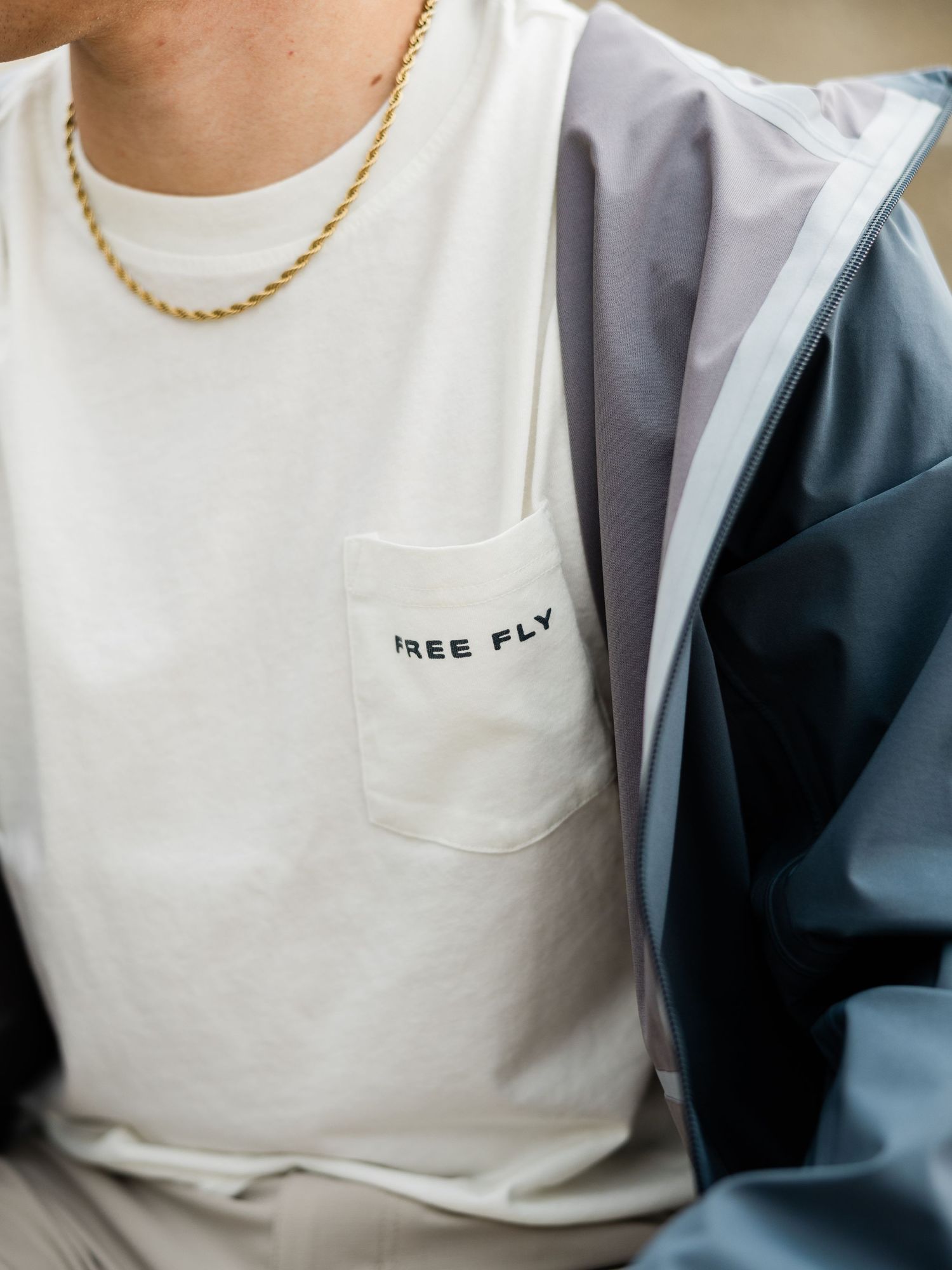 White Free Fly pocket t-shirt worn with a gold chain necklace and blue jacket.