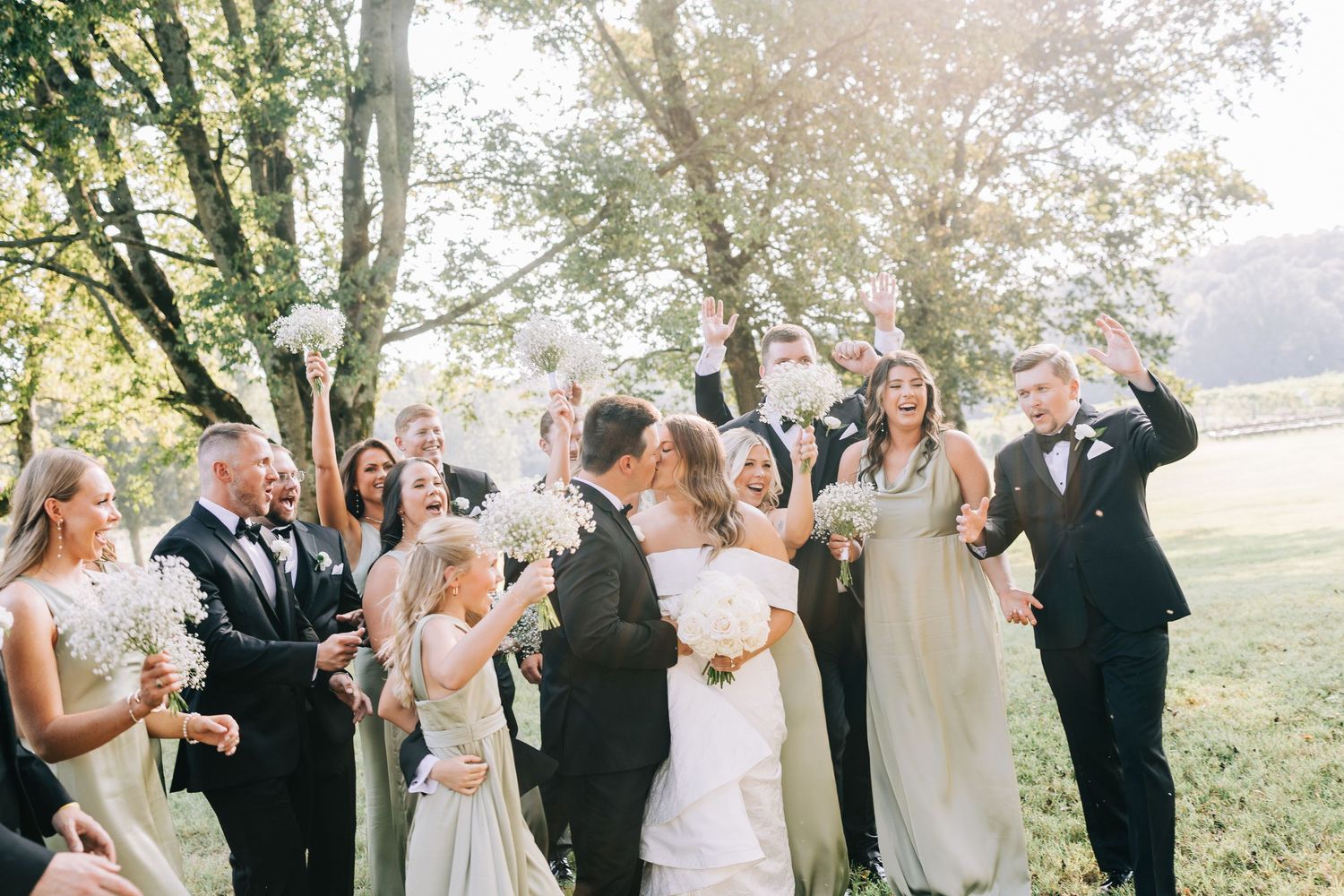 Wedding party celebrates outdoors with joyful dancing and laughter during golden hour in a tree-lined garden setting.