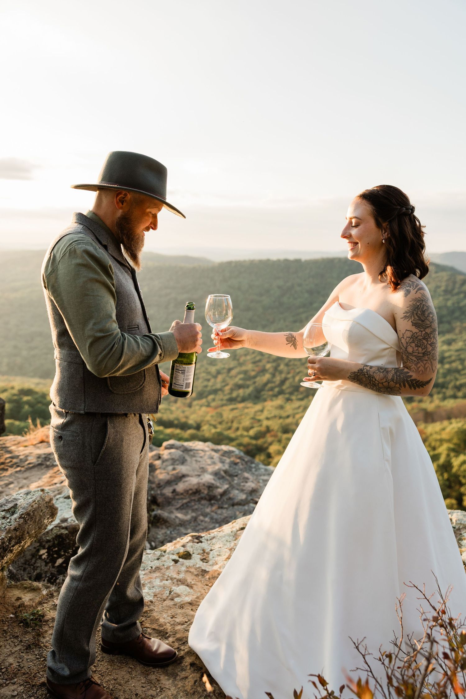 A couple in wedding attire shares drinks while standing on a scenic mountain cliff at sunset.