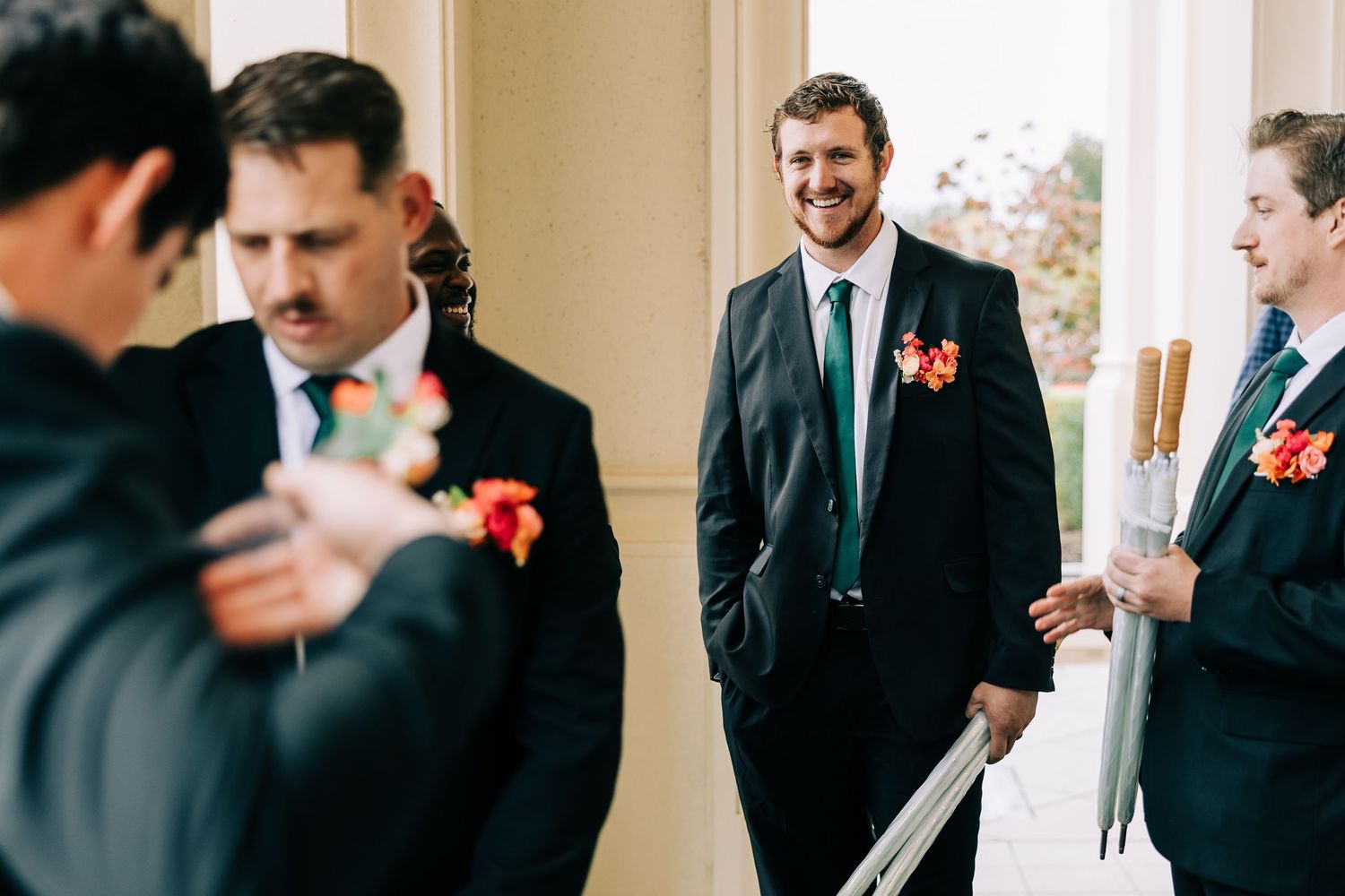 A series of candid wedding moments showing groomsmen in black suits with red boutonnieres laughing in a bright hallway.