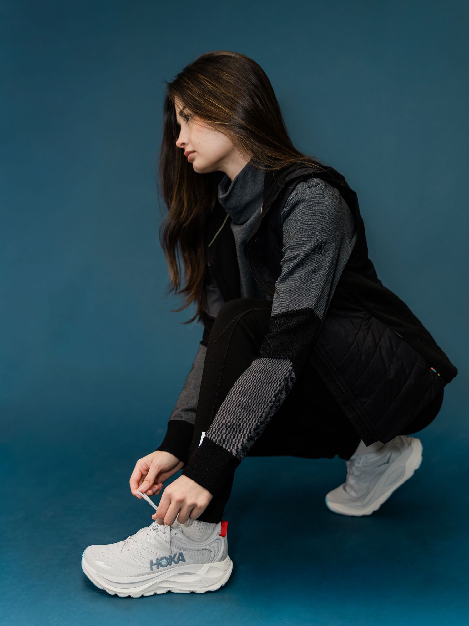 Person in dark clothing bends down to tie white athletic sneakers against a teal studio backdrop.