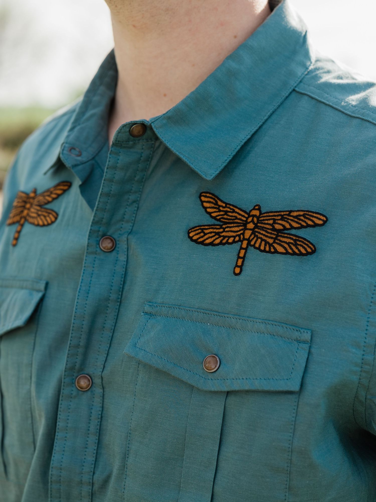 Teal button-up shirt with two embroidered orange dragonfly patches on the chest pockets.