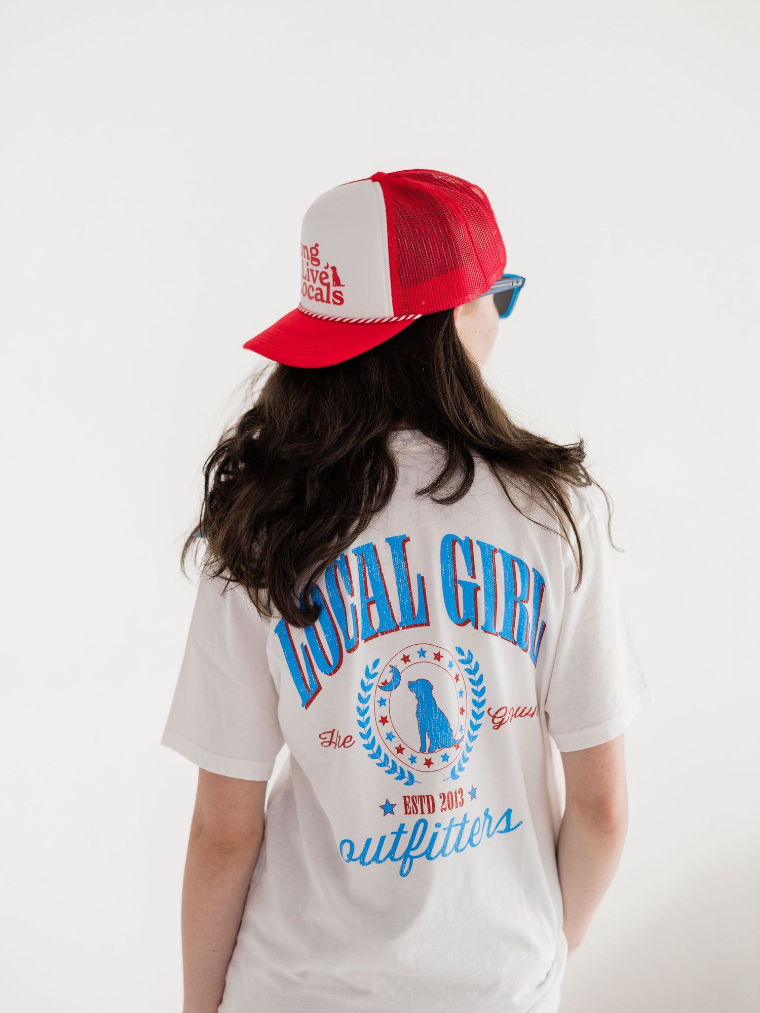 Woman wearing a white Local Girl Outfitters graphic tee and red trucker hat, shown from behind.