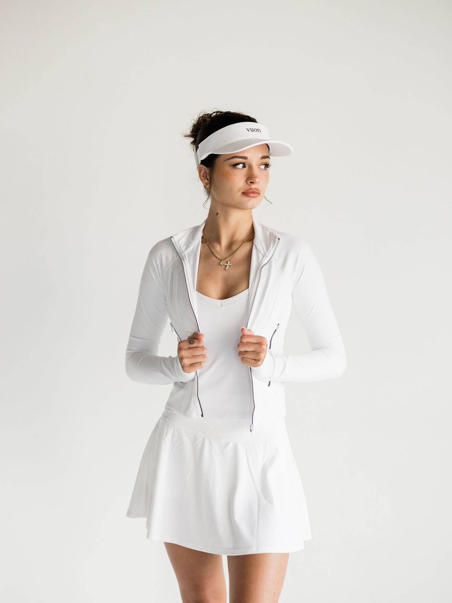 Woman wearing white athletic outfit with zip-up jacket, skirt, and visor, posing against a light background.