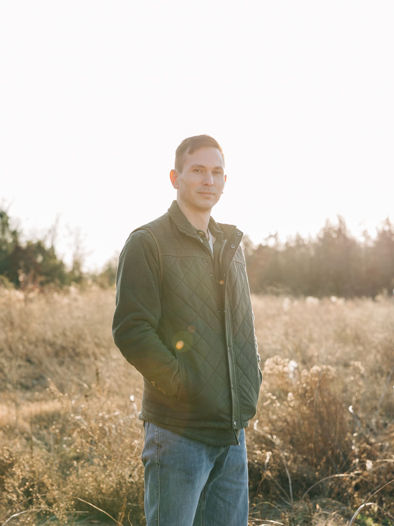 Young man in quilted jacket and jeans standing in golden field at sunset with warm backlight.