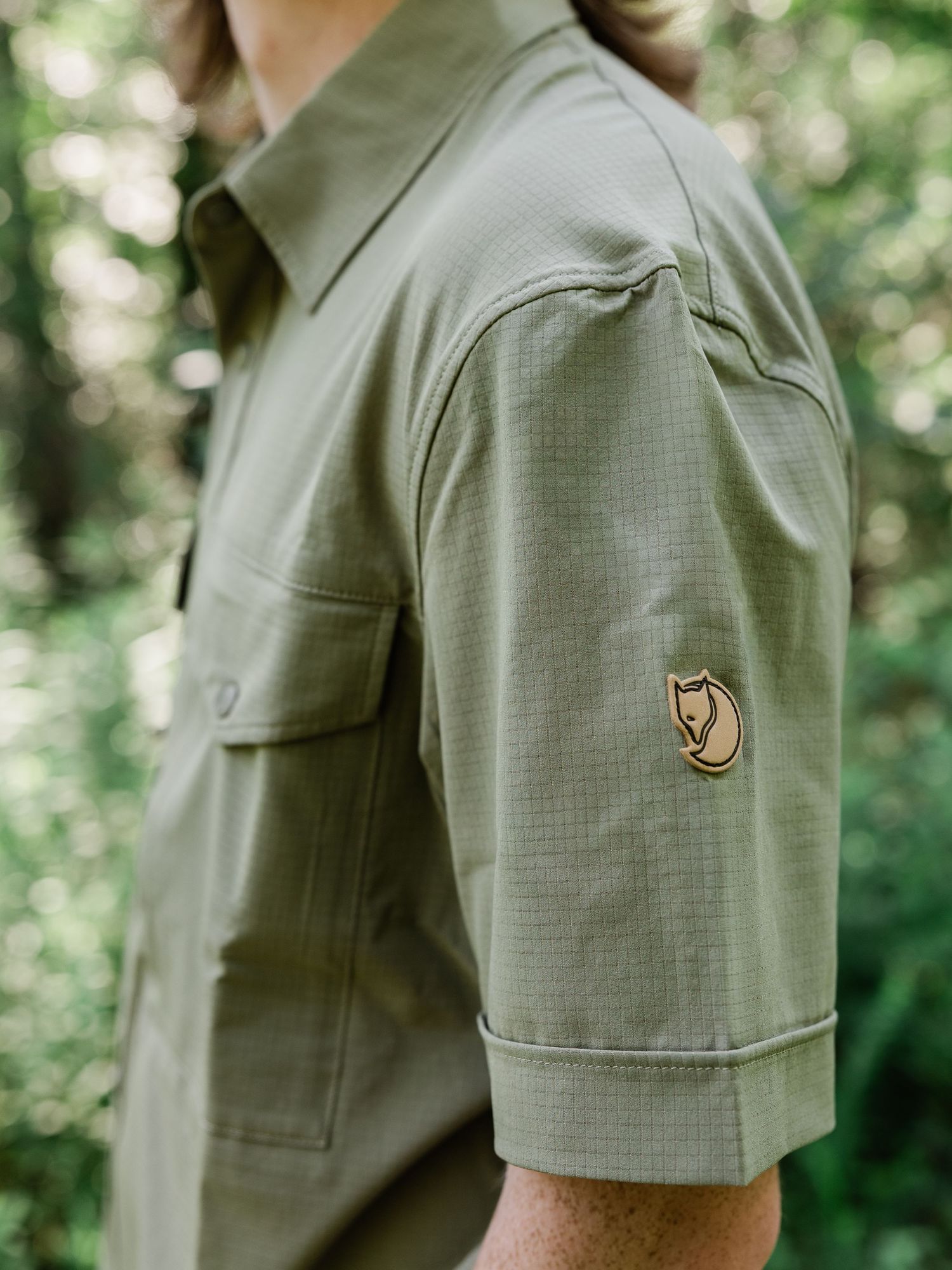 Fjallraven olive green short-sleeve shirt with fox logo patch, worn outdoors in a forest setting.