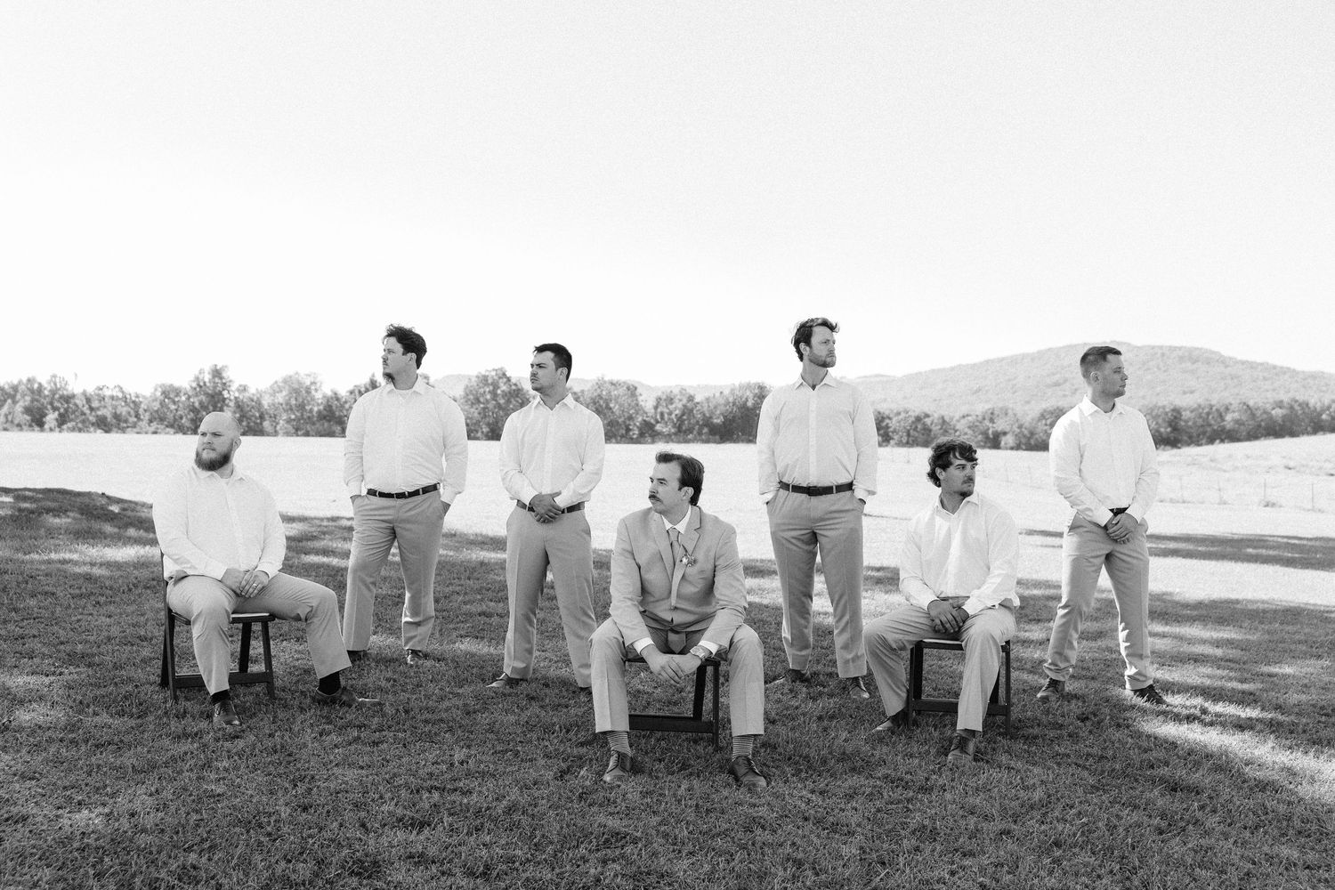 Wedding party members seated in white chairs on a grassy hill for group photos.