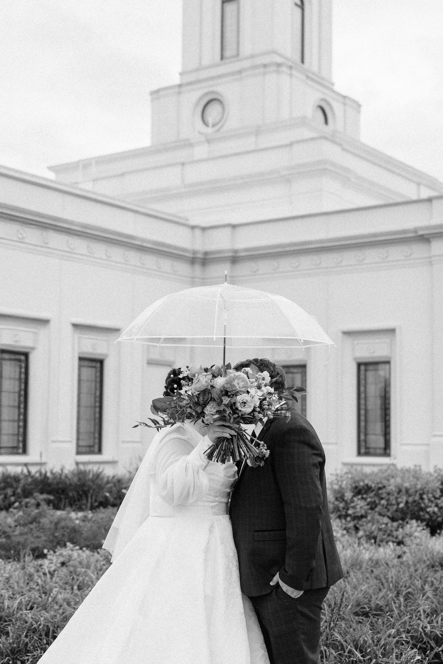 Wedding couple poses with clear umbrella in front of white church architecture.