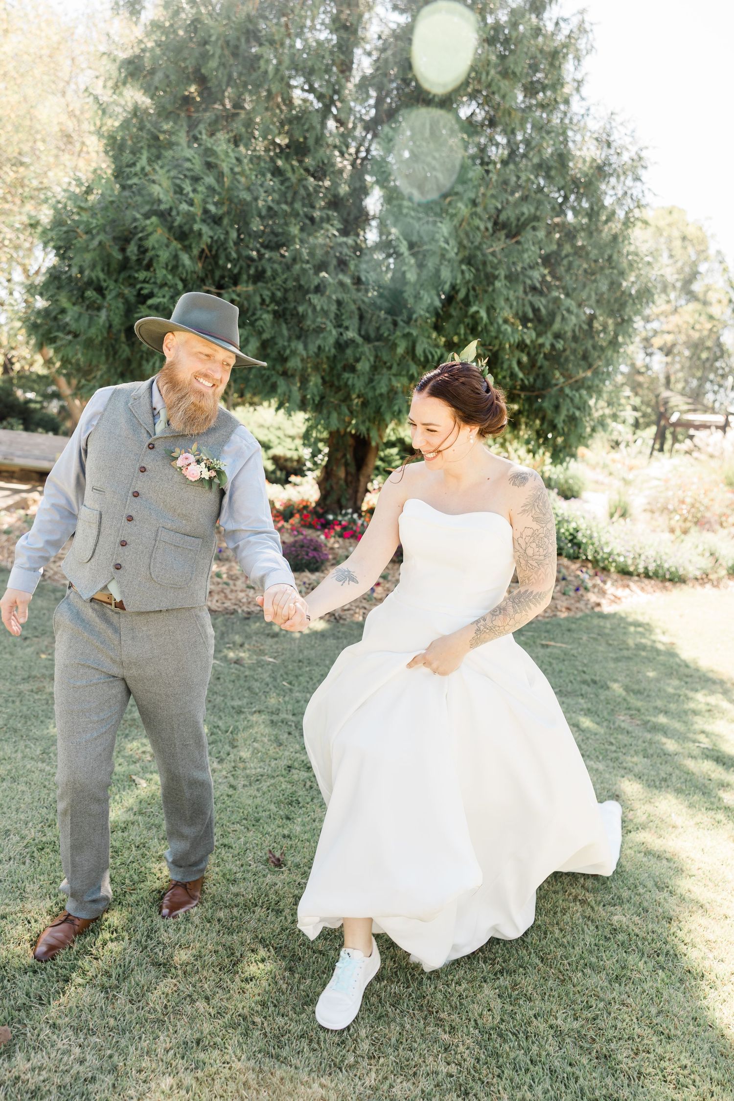 A couple in wedding attire playfully dances together outdoors surrounded by greenery on a sunny day.