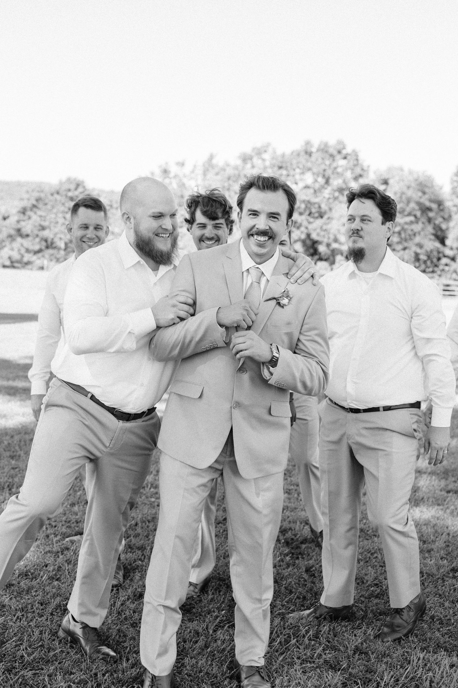 Groomsmen in light colored suits share laughs during outdoor wedding photo session.