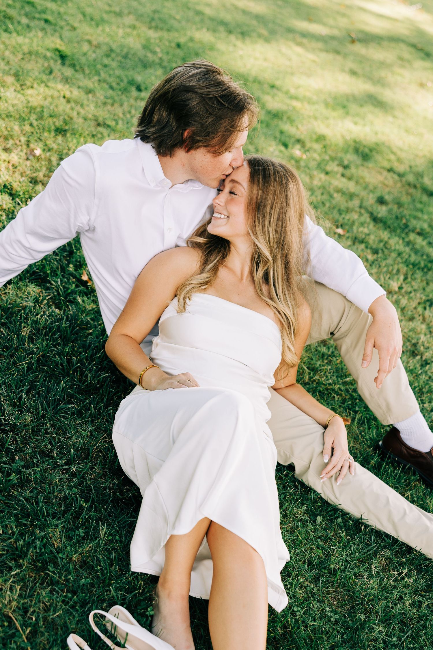 Romantic couple in white outfits embracing on green grass during a sunny outdoor photoshoot.