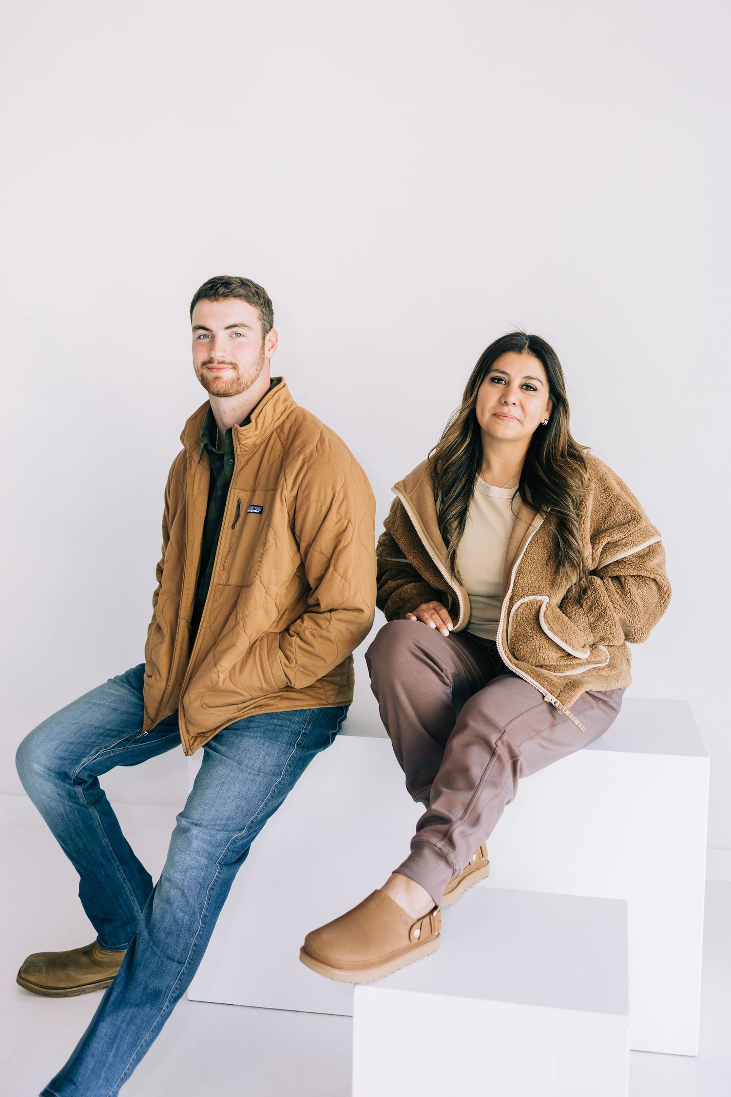 Two models sitting on white cube wearing tan sherpa jackets and UGG-style boots in a minimalist studio setting.