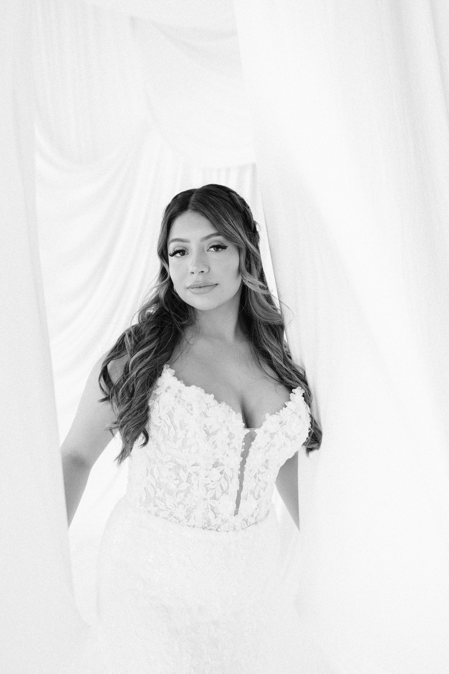 Bride wearing delicate white lace wedding gown poses elegantly against light background.