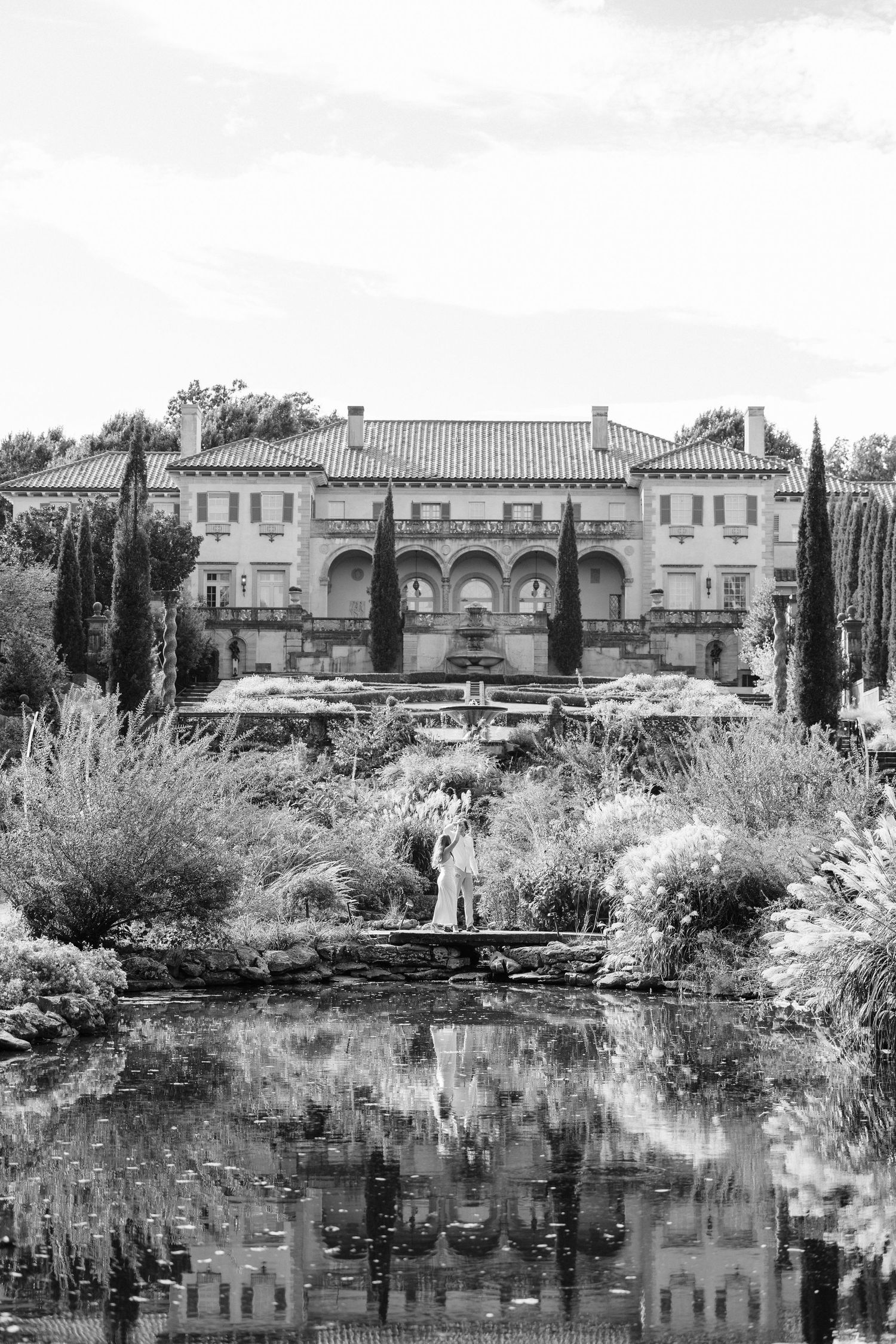 Black and white panoramic view of a serene garden pond with ornate buildings and cypress trees reflected in the water.