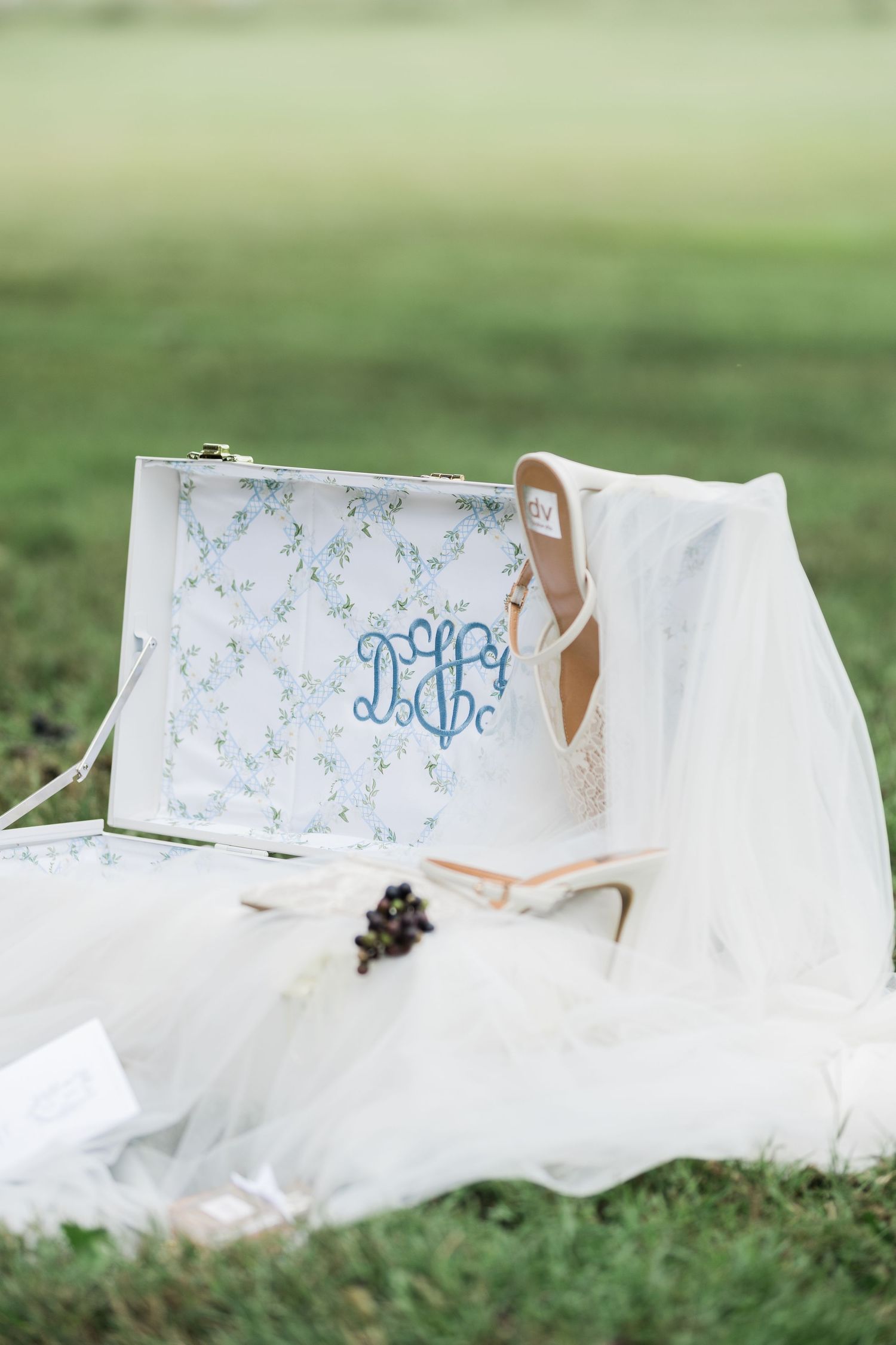 White bridal accessories including monogrammed clutch, delicate veil and elegant heels arranged on grass outdoors.