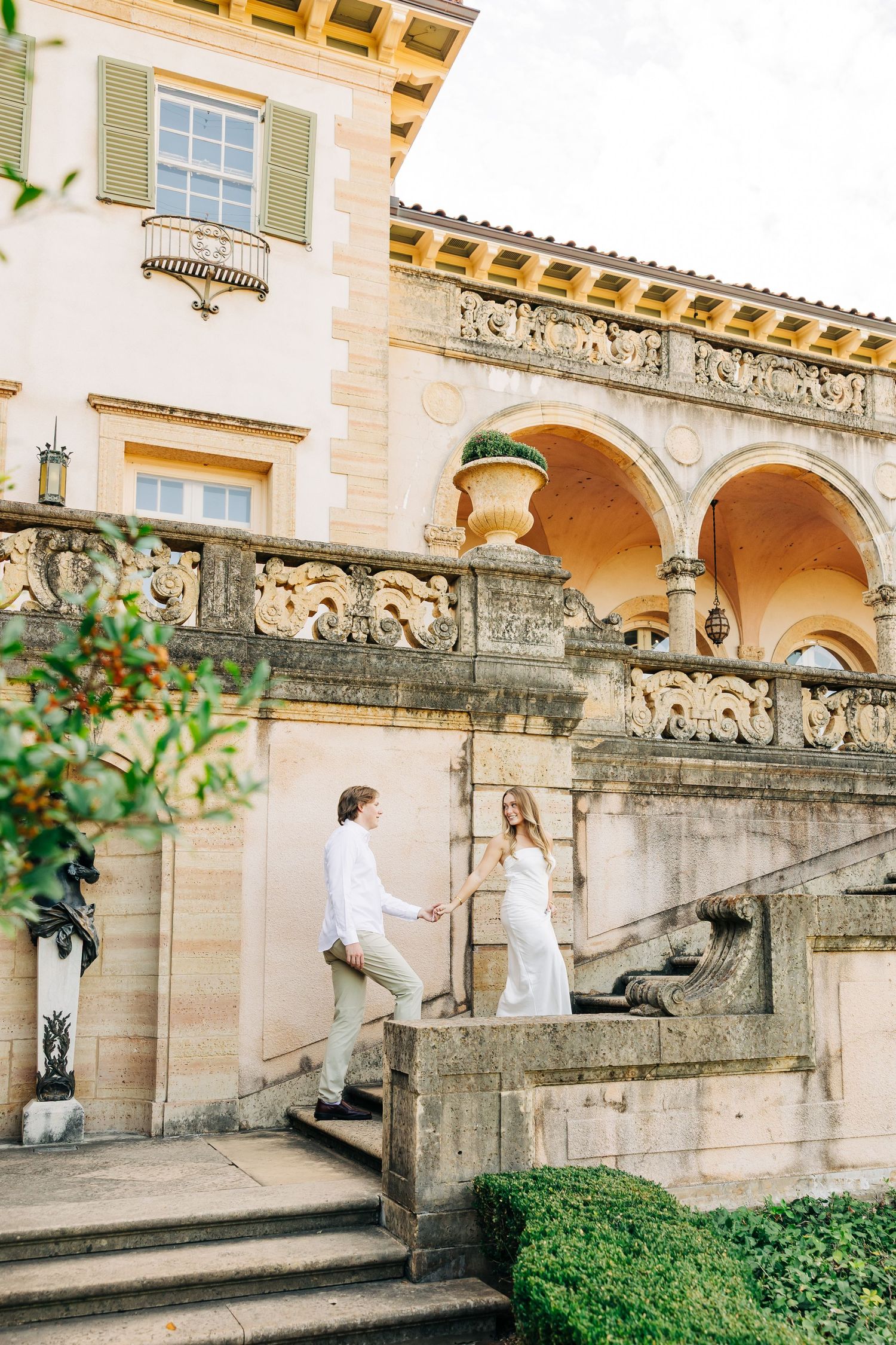 Elegant couple descends stone steps at a Mediterranean-style villa with arched columns and lush greenery.