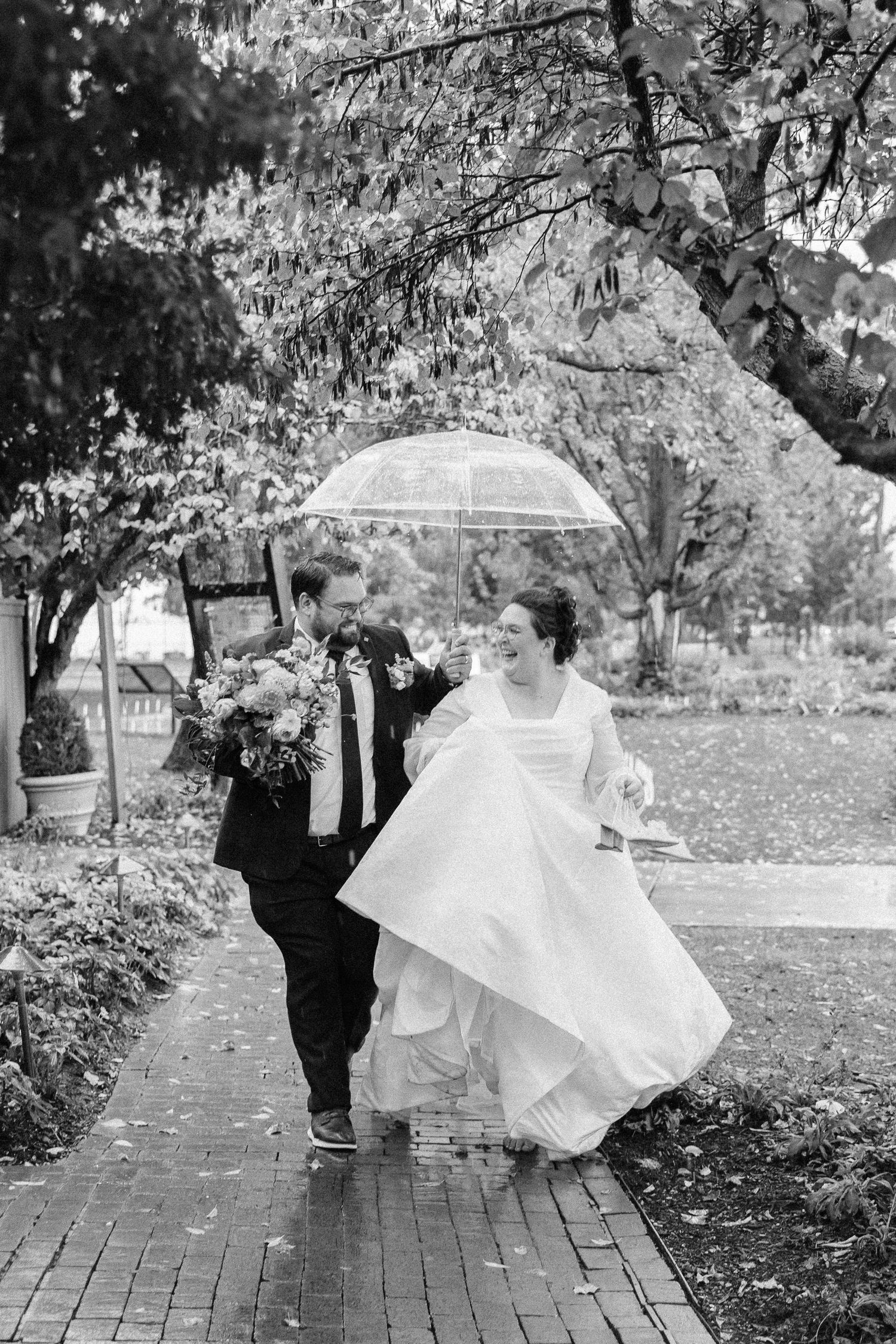 A couple walks along a brick path under an umbrella on their rainy wedding day.