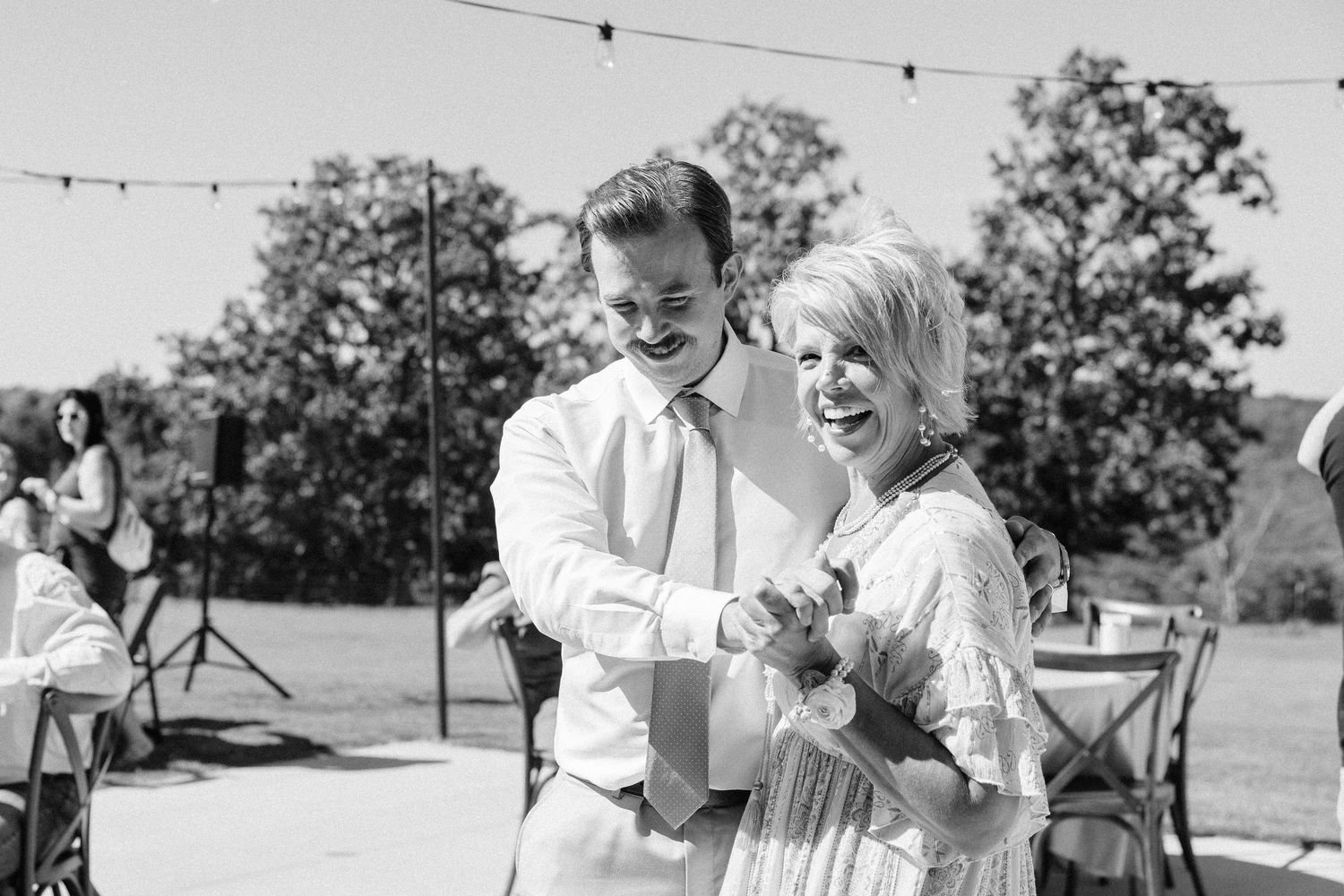 Black and white photo of guests mingling at an outdoor celebration with string lights overhead.