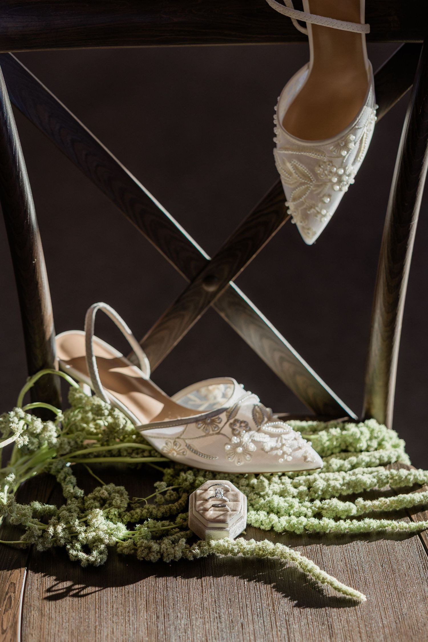Elegant white bridal shoes with pearl details displayed on a wooden ledge with green floral accents.