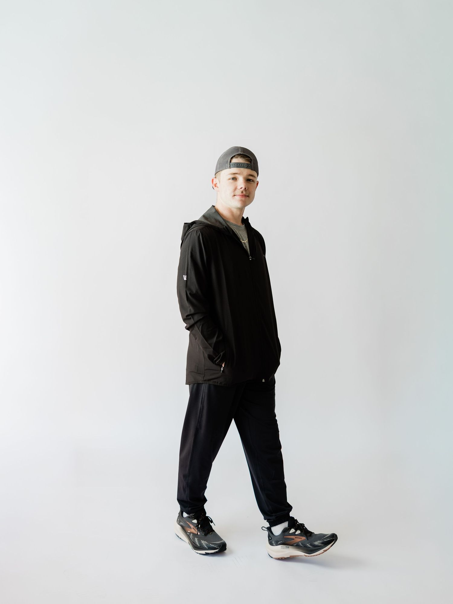 Person wearing black athletic wear and sneakers poses against white background in minimalist studio setting.