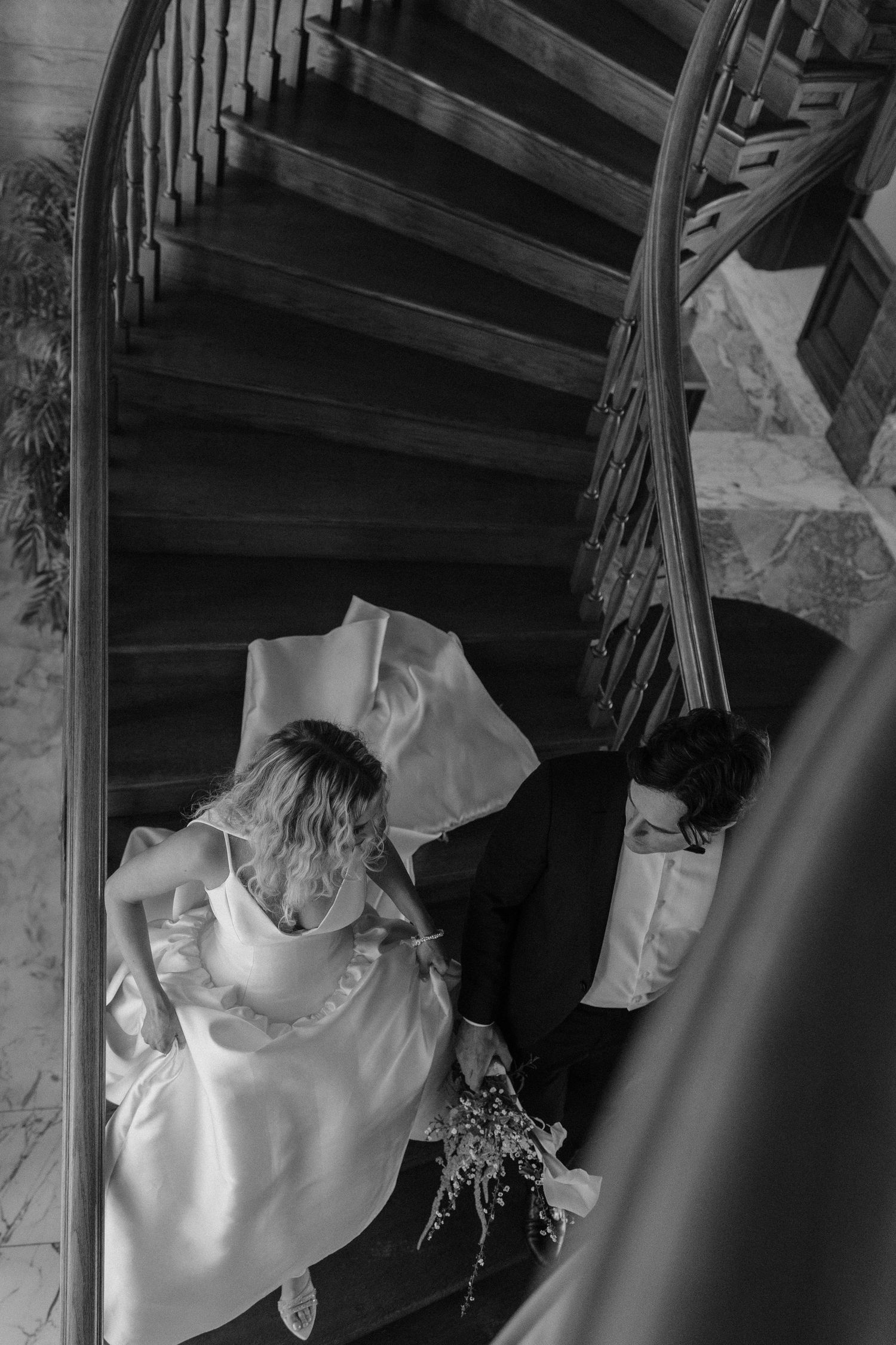A couple descends a curved wooden staircase in formal attire, with the person in a white wedding dress holding a bouquet.