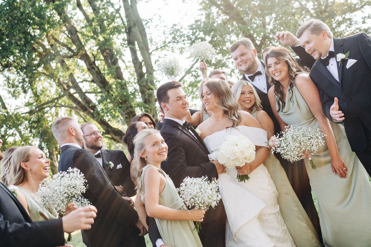 Wedding party laughs together outdoors holding white bouquets in formal attire under sunlit trees.