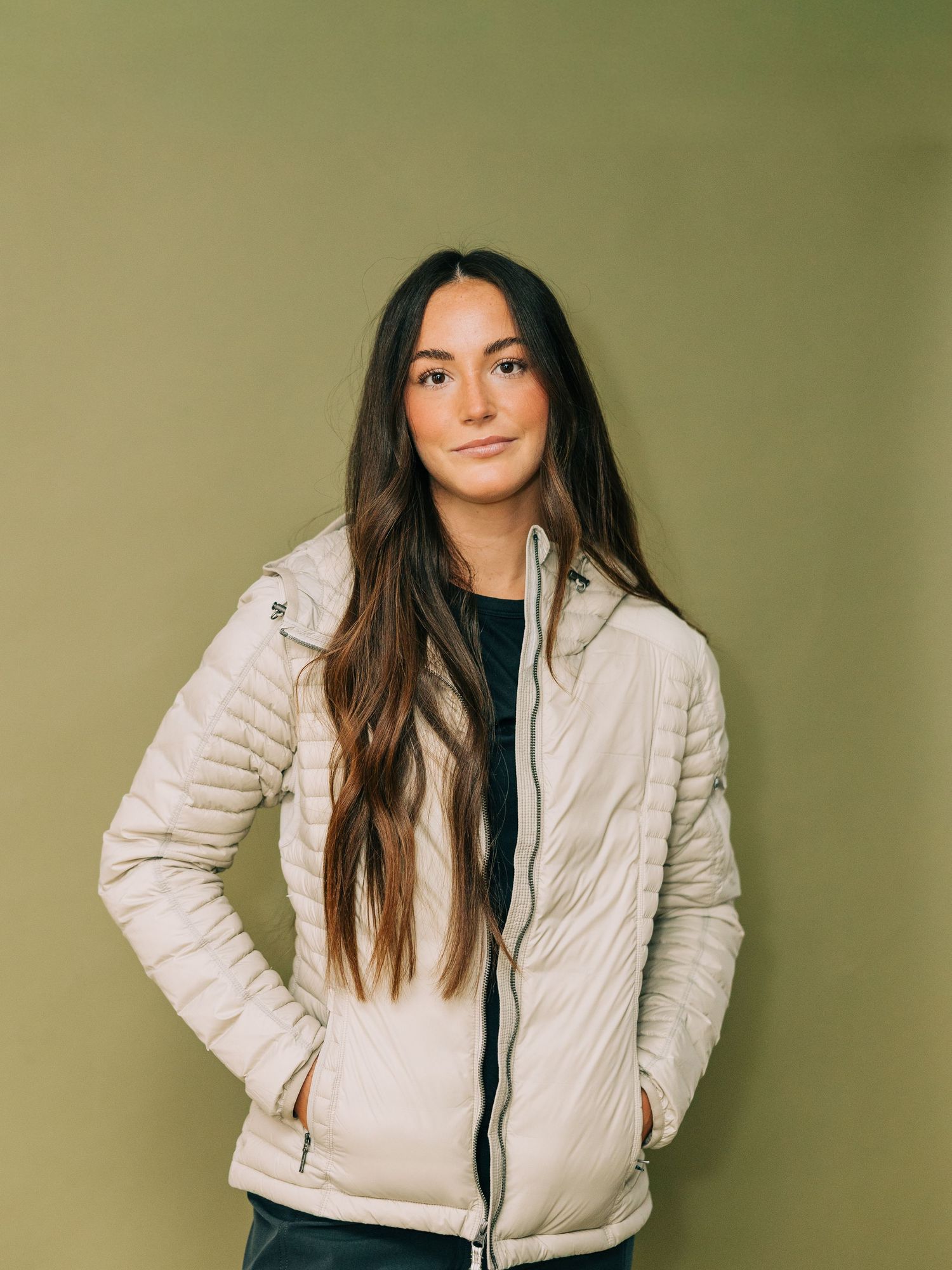 A person wearing a cream colored puffer jacket and long dark hair poses against an olive green background.