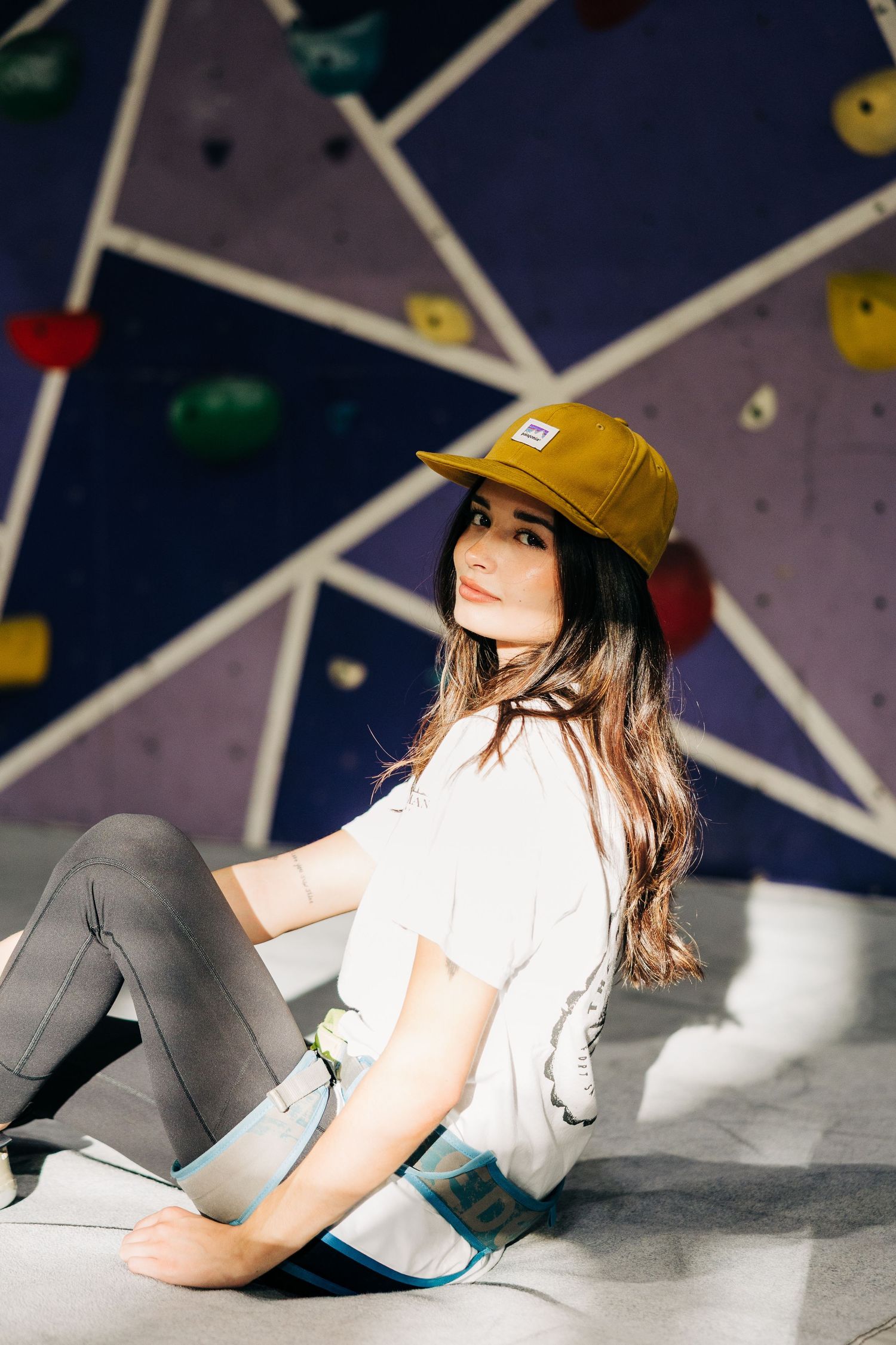 A person in a yellow baseball cap and white top poses on a climbing wall with geometric patterns in the background.