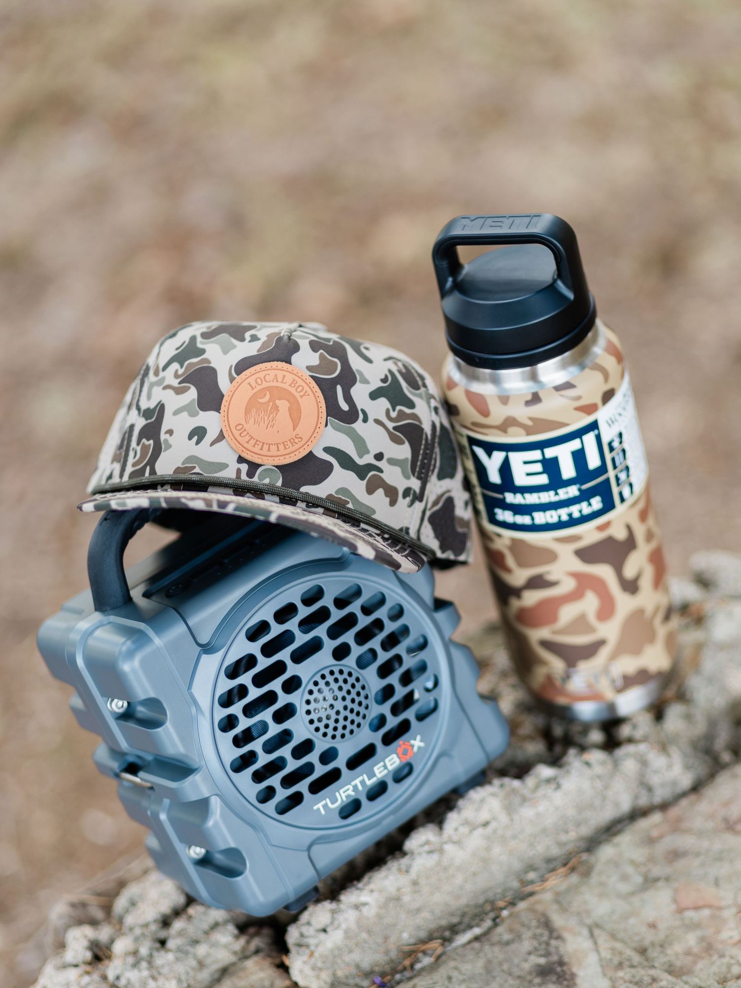 Camo YETI Rambler bottle, TurtleBox speaker, and camo hat arranged on a rock outdoors.
