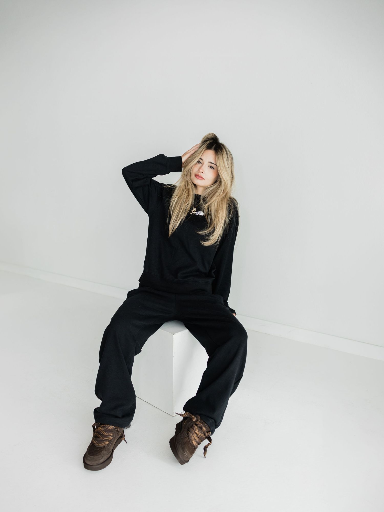Person in black oversized hoodie and wide-leg pants with brown boots, seated casually on white cube against light background.