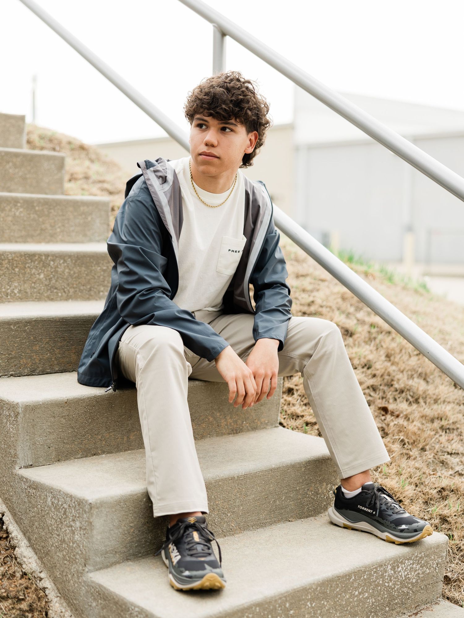 Teen boy in navy jacket and khaki pants sitting on concrete steps wearing dark sneakers with a backpack.
