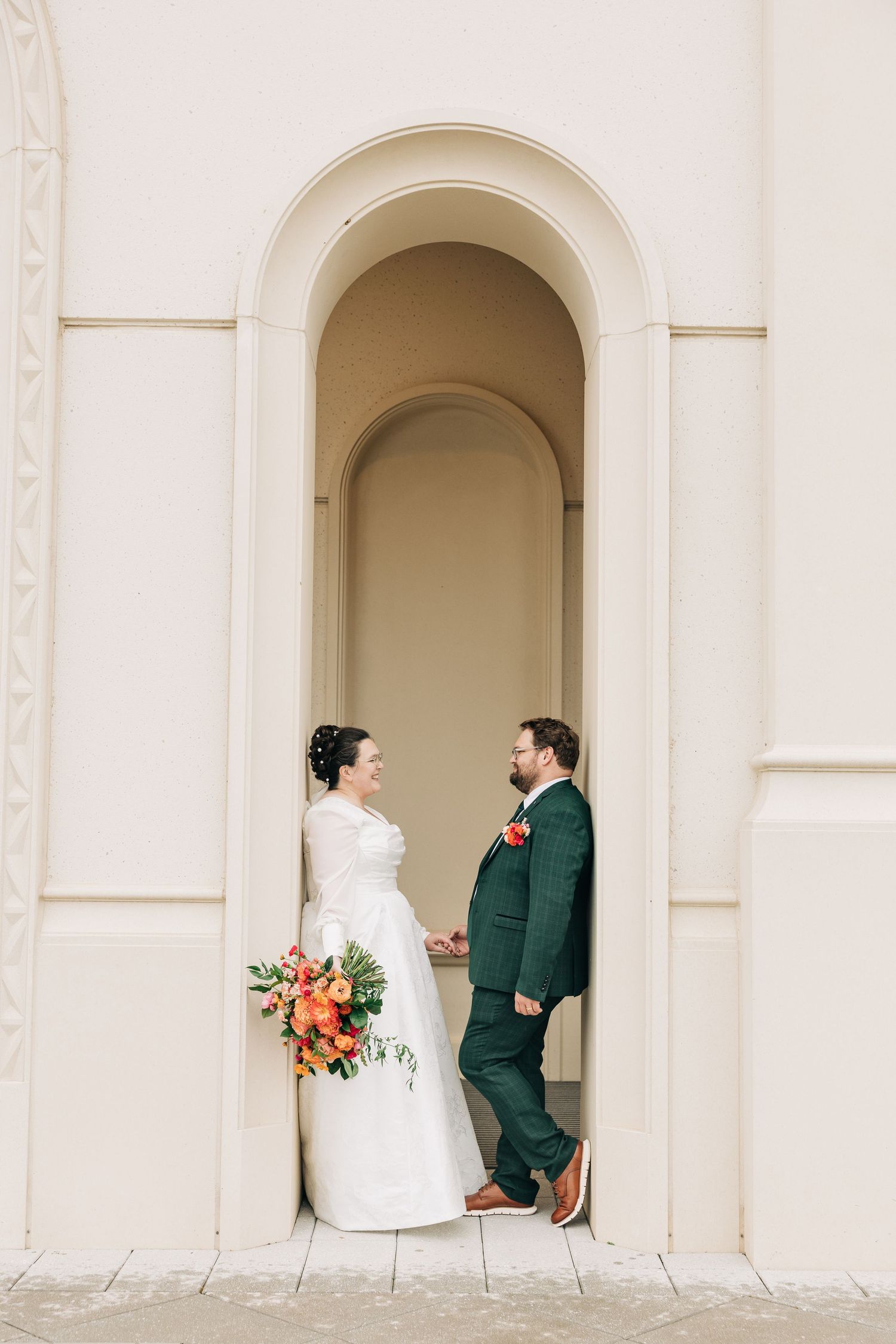 Wedding couple posing in arched alcoves of white church exterior.