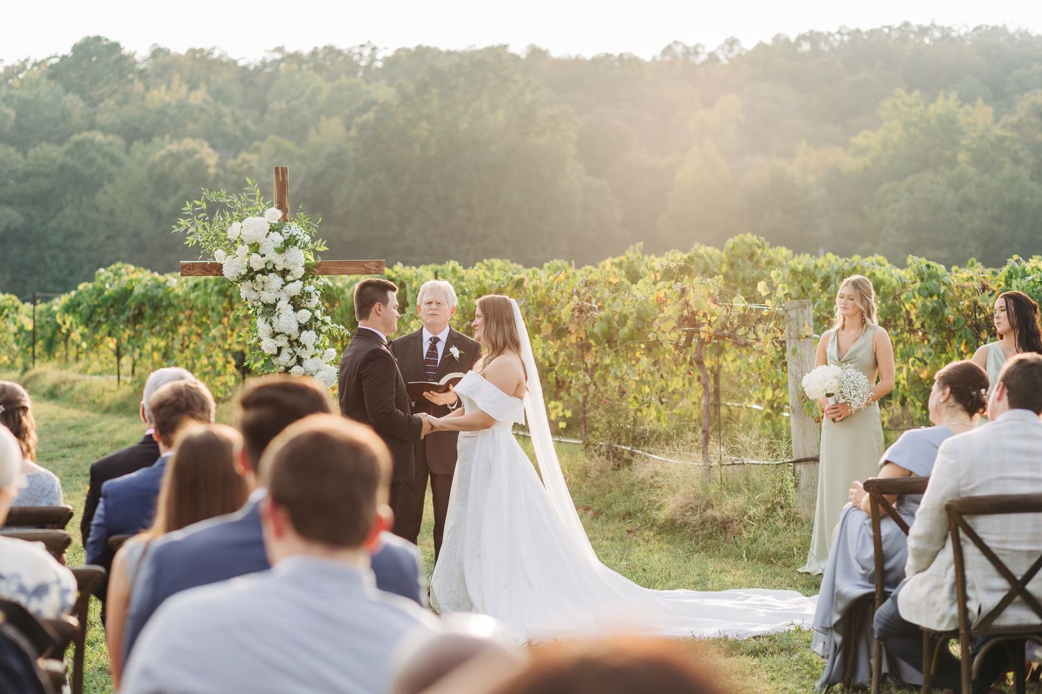 Outdoor vineyard wedding ceremony at sunset with guests seated in white chairs among grapevines.