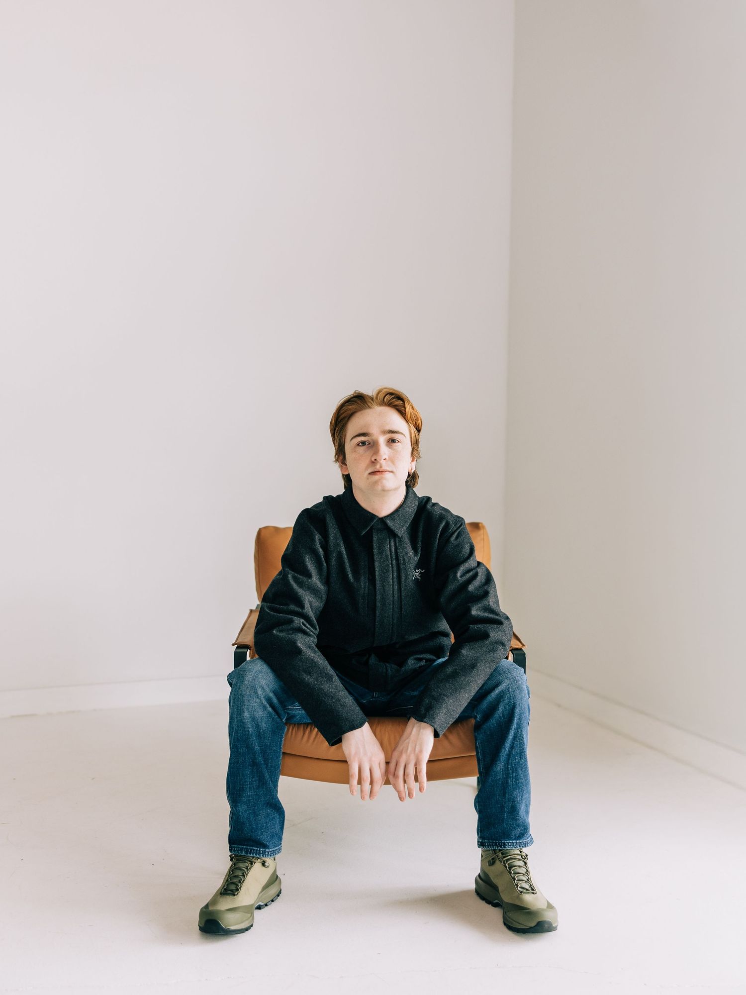 Person in black hoodie and jeans sits with legs spread wide apart on white studio floor.