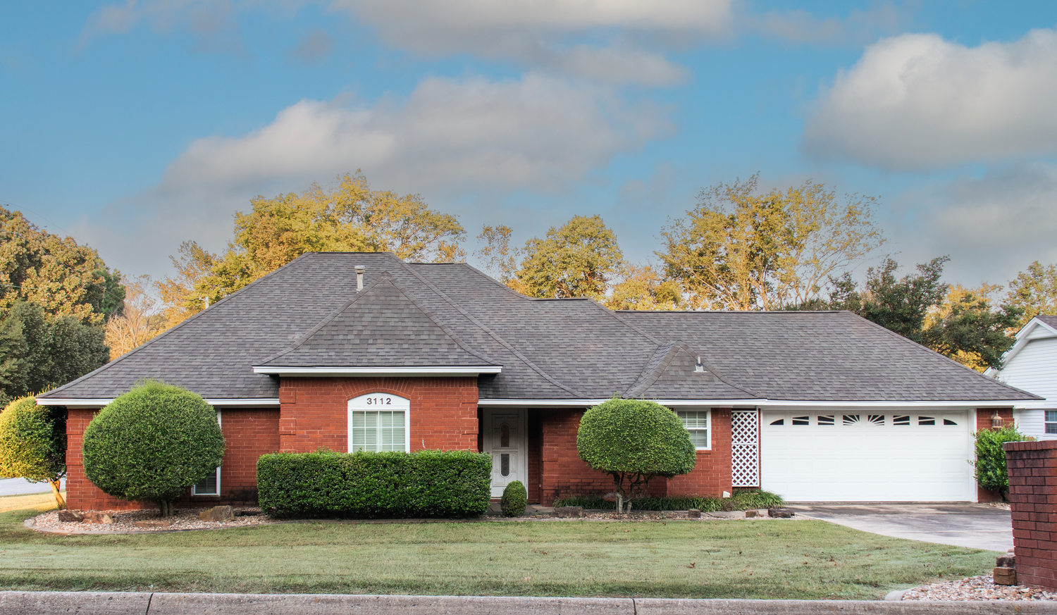 Brick ranch-style home with gray roof, white garage, manicured shrubs, and autumn trees under a partly cloudy sky.
