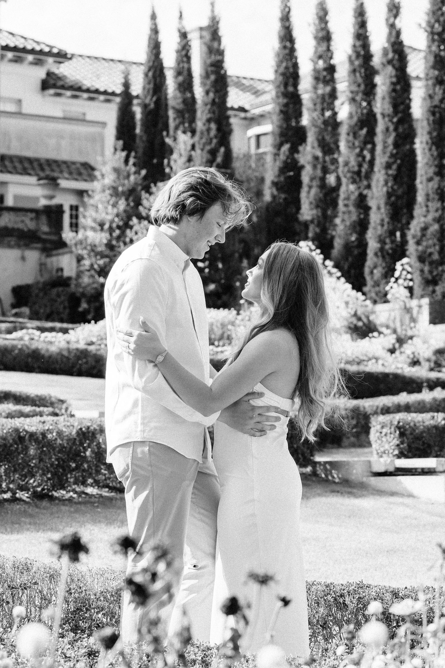 Black and white romantic garden engagement session with cypress trees and manicured hedges in the background.