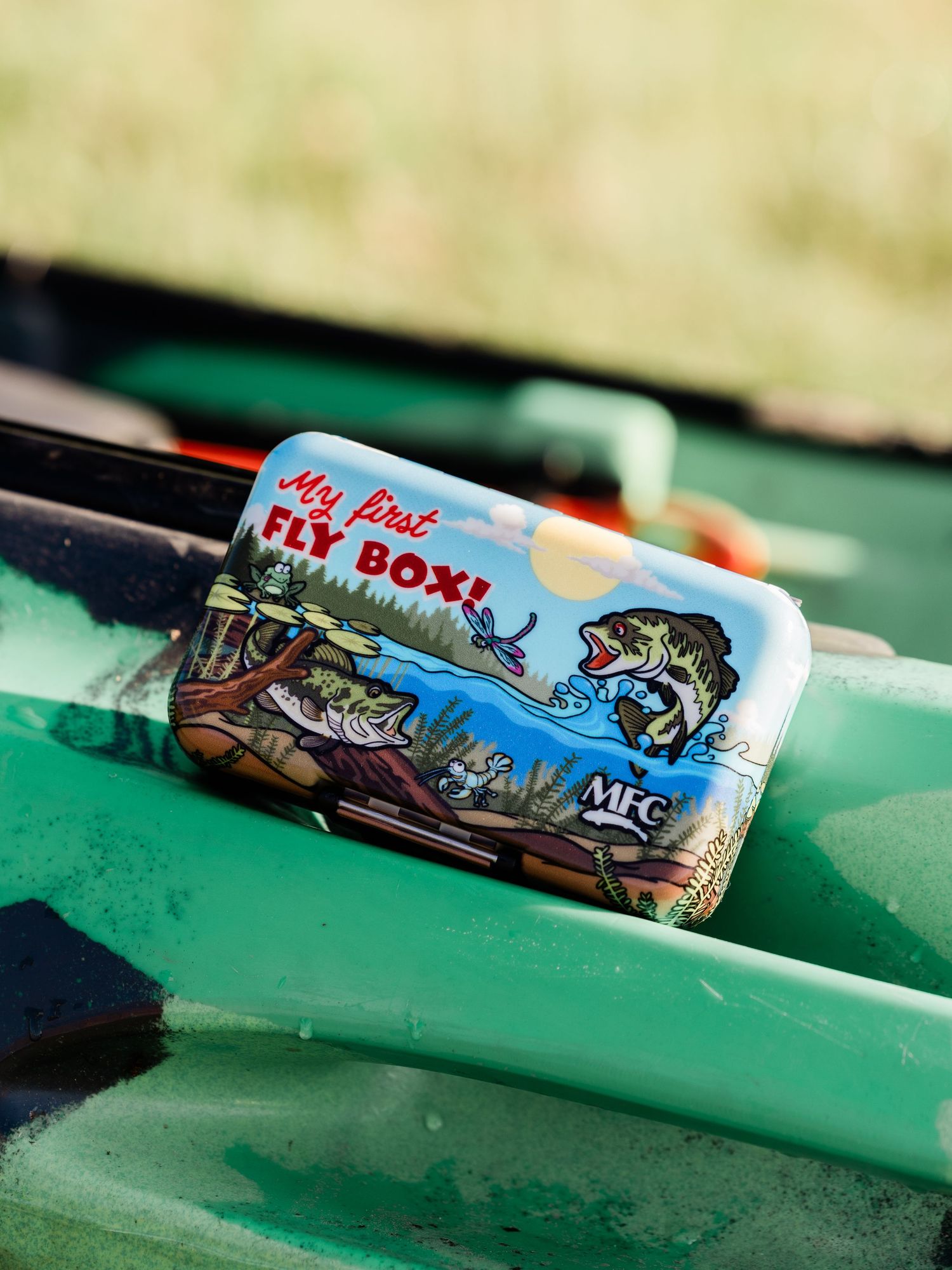 MFC My First Fly Box colorful tin case resting on green fishing tackle box outdoors.