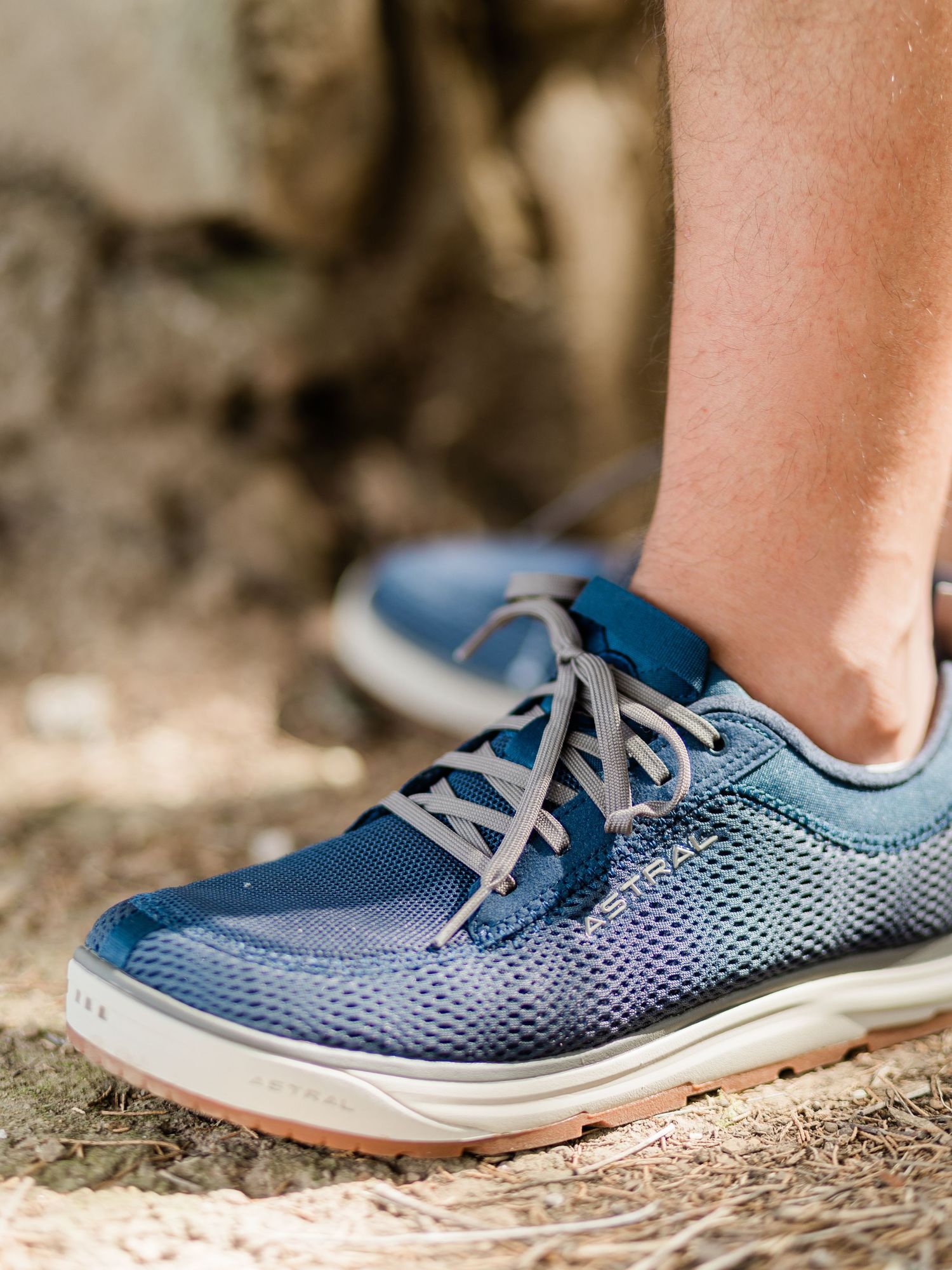 Close-up of a person wearing blue mesh lace-up sneakers with gray laces and white soles on a natural ground.